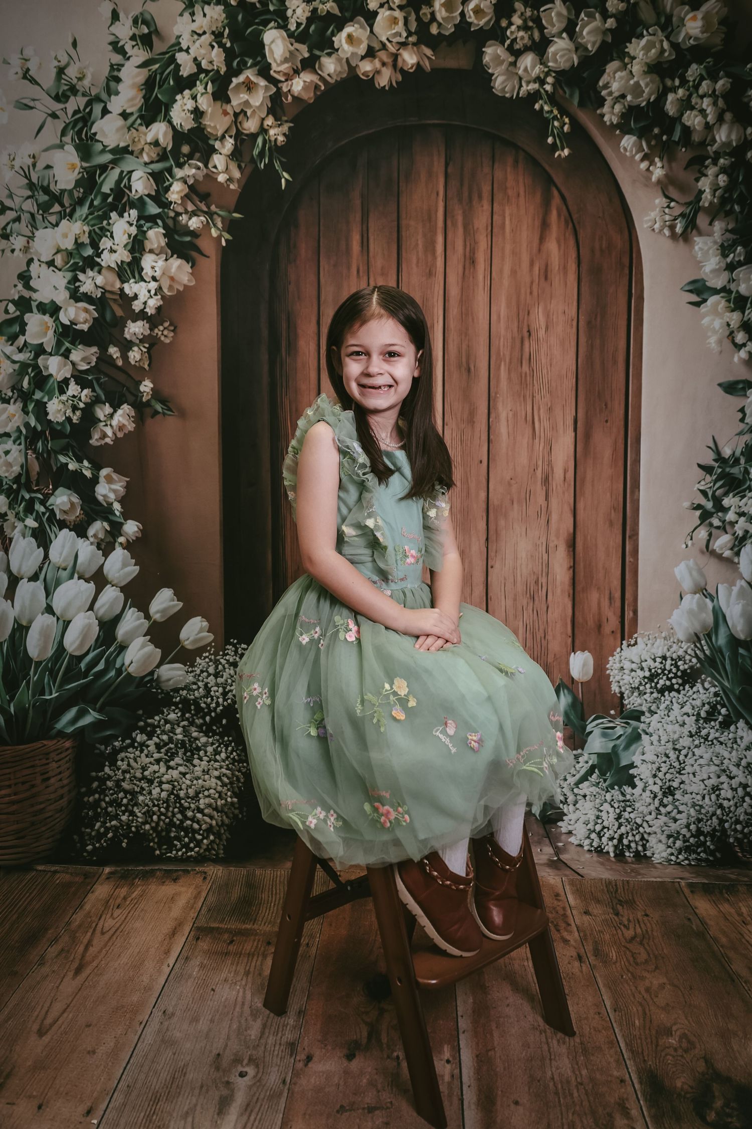 Young girl in green floral dress sits on stool before wooden arch door surrounded by white tulips and roses.