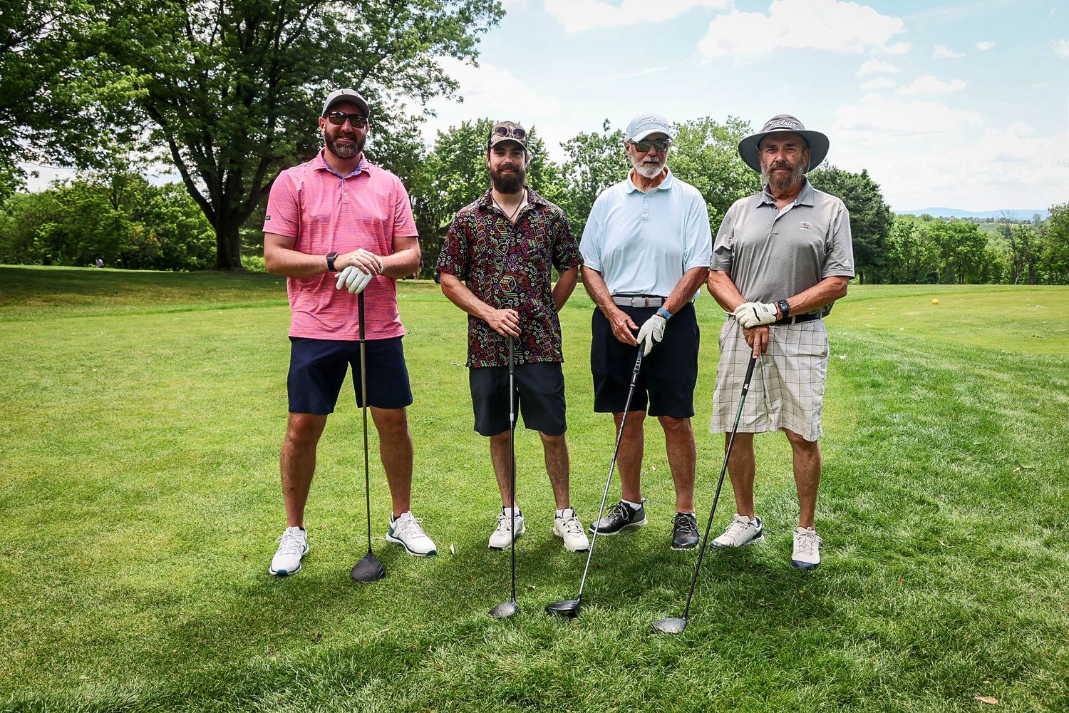 Four men posing with golf clubs on a lush green golf course on a sunny day.