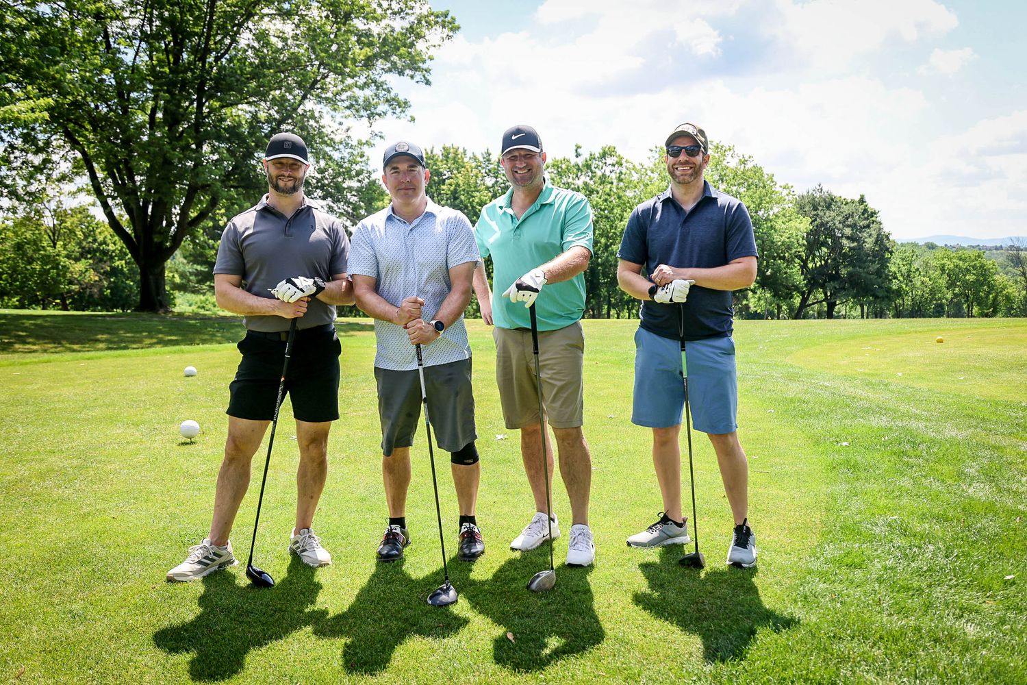 Four men pose with golf clubs on a sunny green golf course on a clear summer day.
