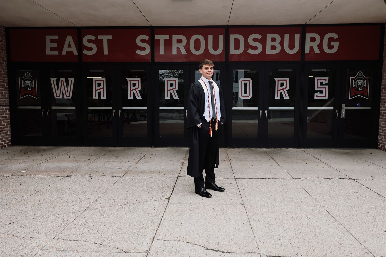 Graduate in cap and gown stands proudly in front of East Stroudsburg Warriors school entrance.