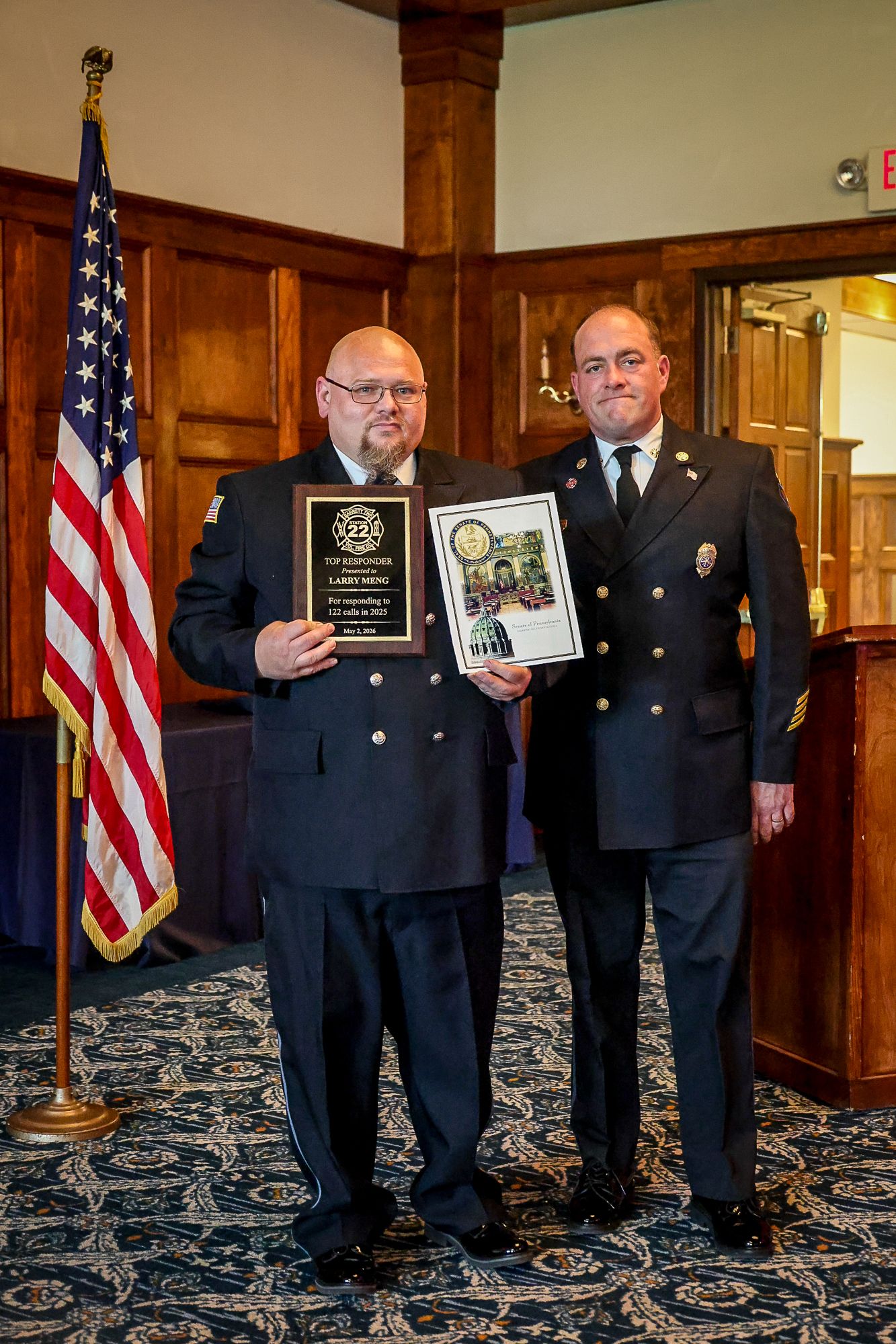 Two uniformed firefighters posing with award plaques at a formal ceremony, American flag displayed in background.
