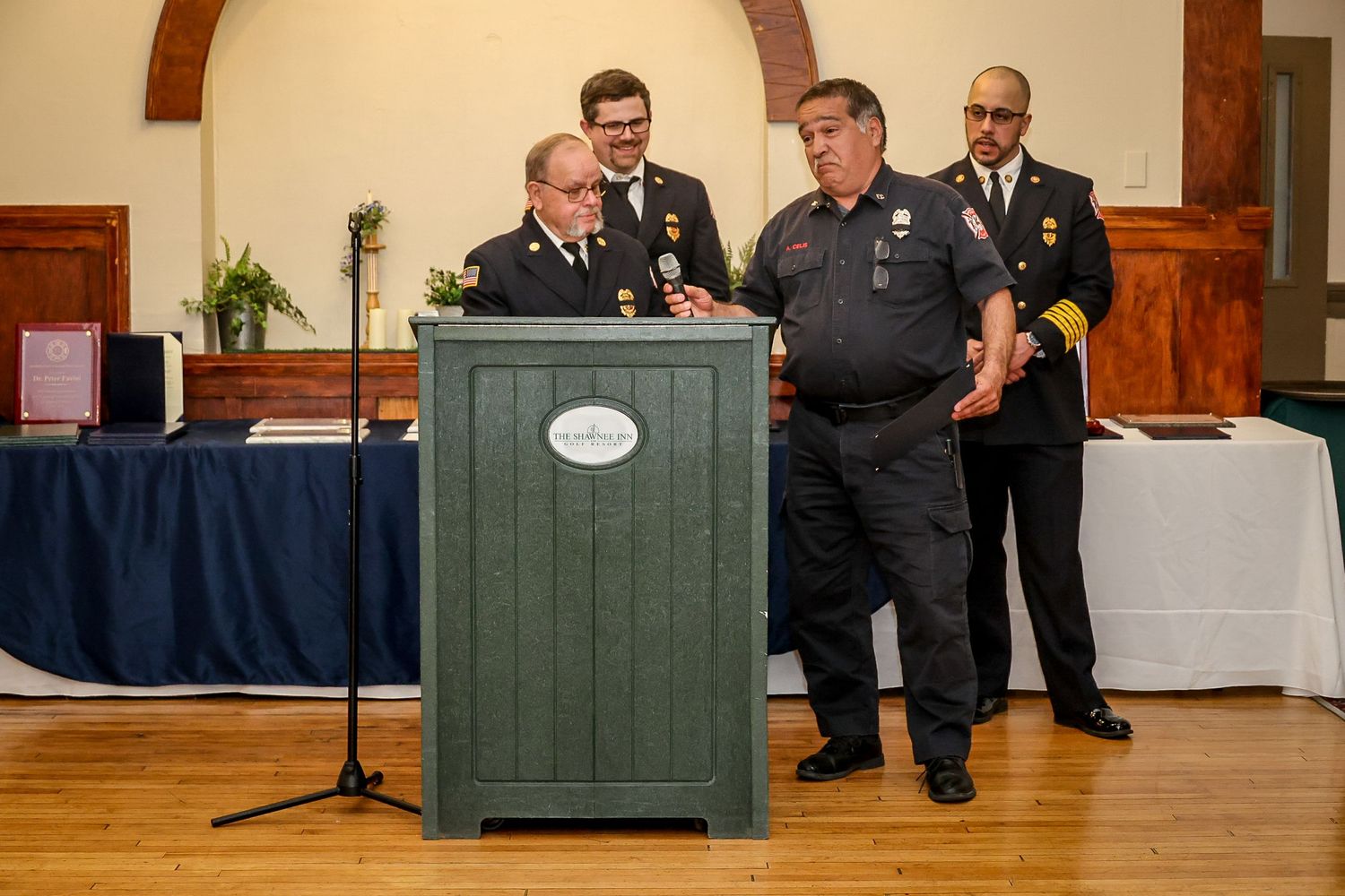 Firefighters in dress uniforms gather at a green podium during an awards ceremony in a decorated hall.