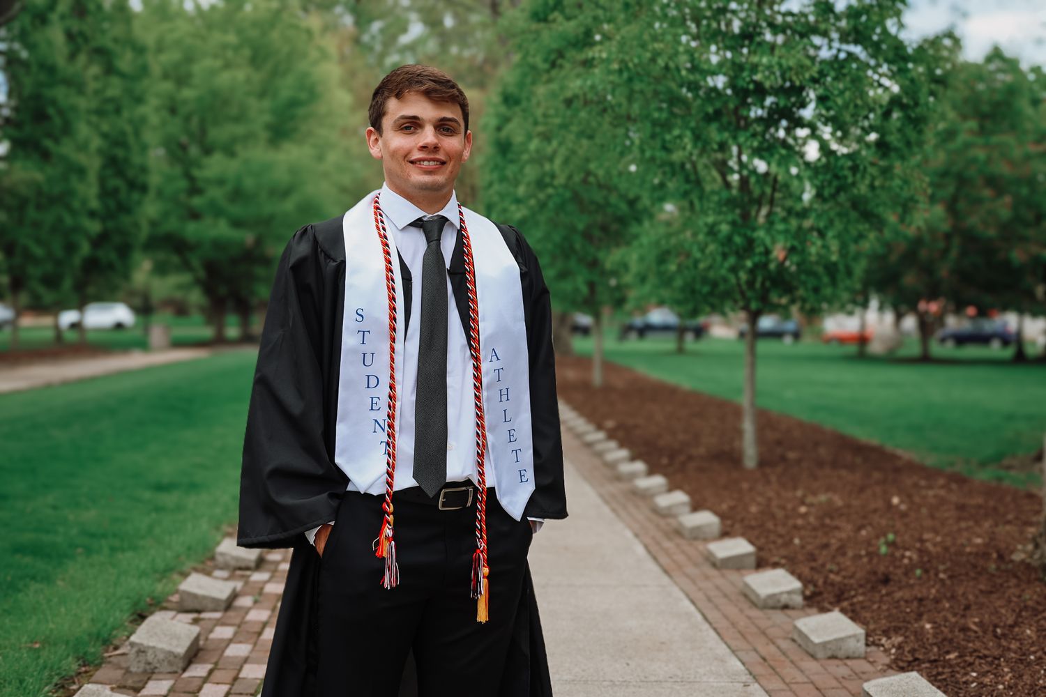 Young man in graduation gown and stole smiling on a tree-lined campus walkway on graduation day.