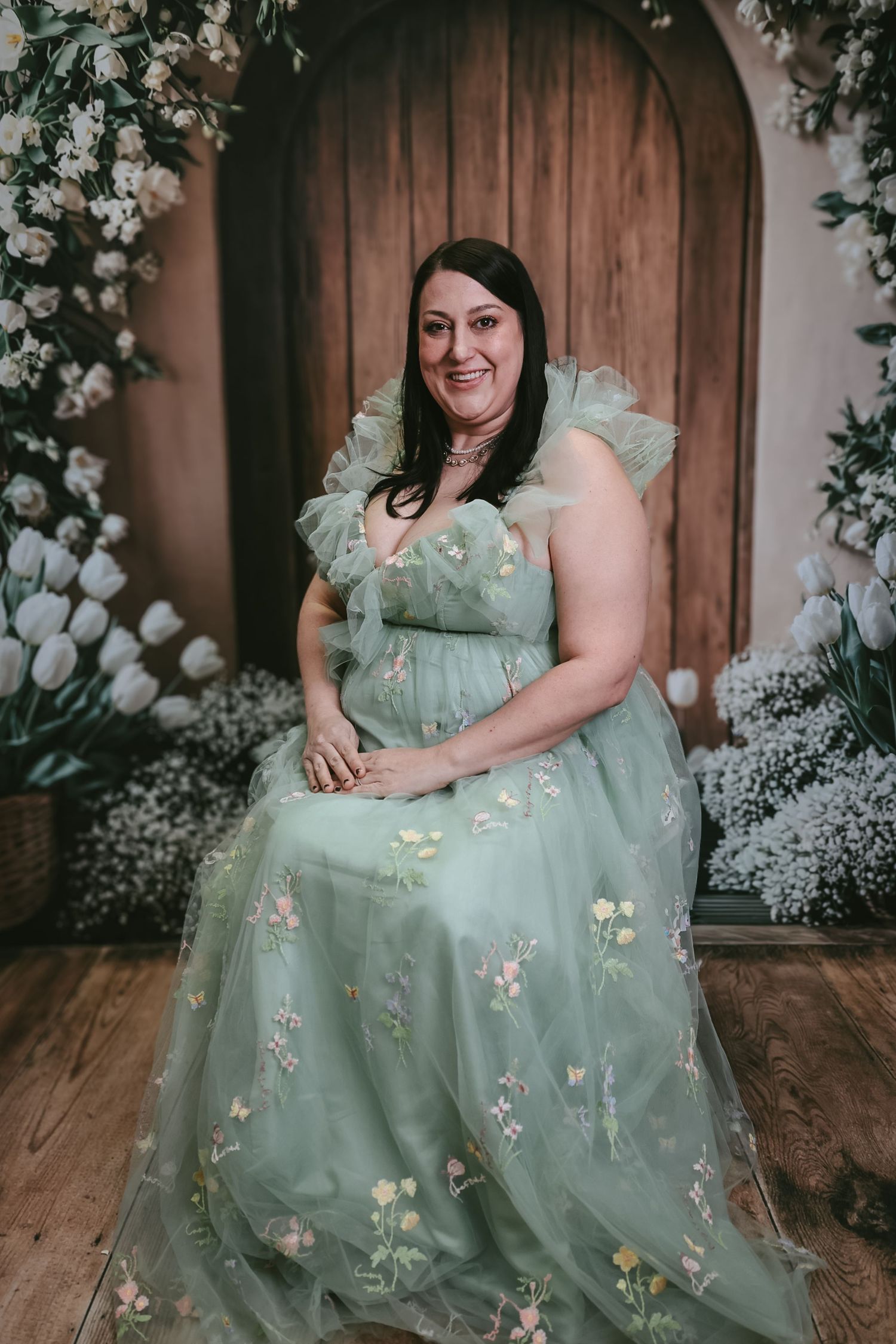 Woman in sage green floral tulle gown sits smiling before a floral backdrop with white tulips.