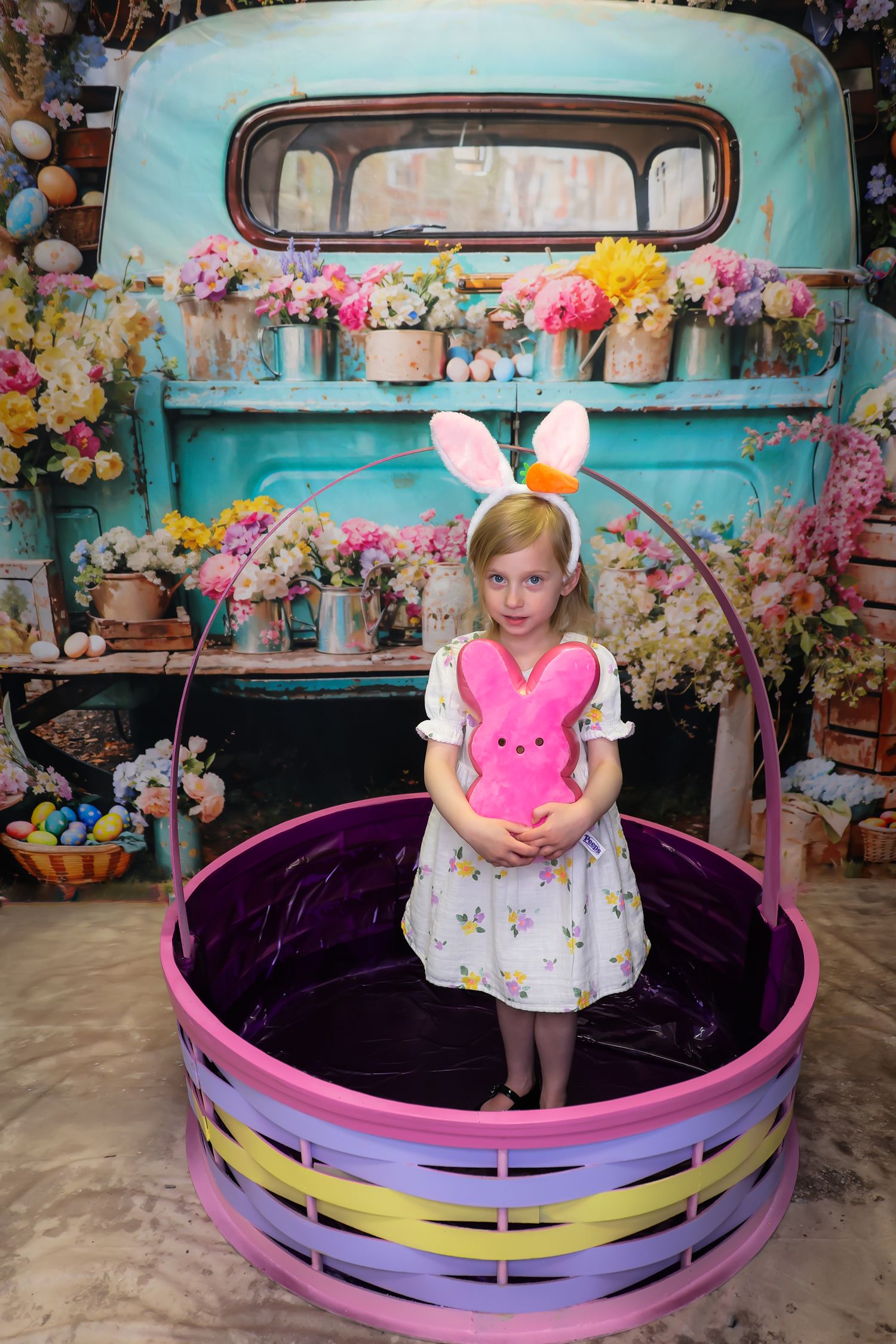 Young girl in bunny ears holds pink Peeps bunny in colorful Easter basket, vintage teal truck with flowers backdrop.
