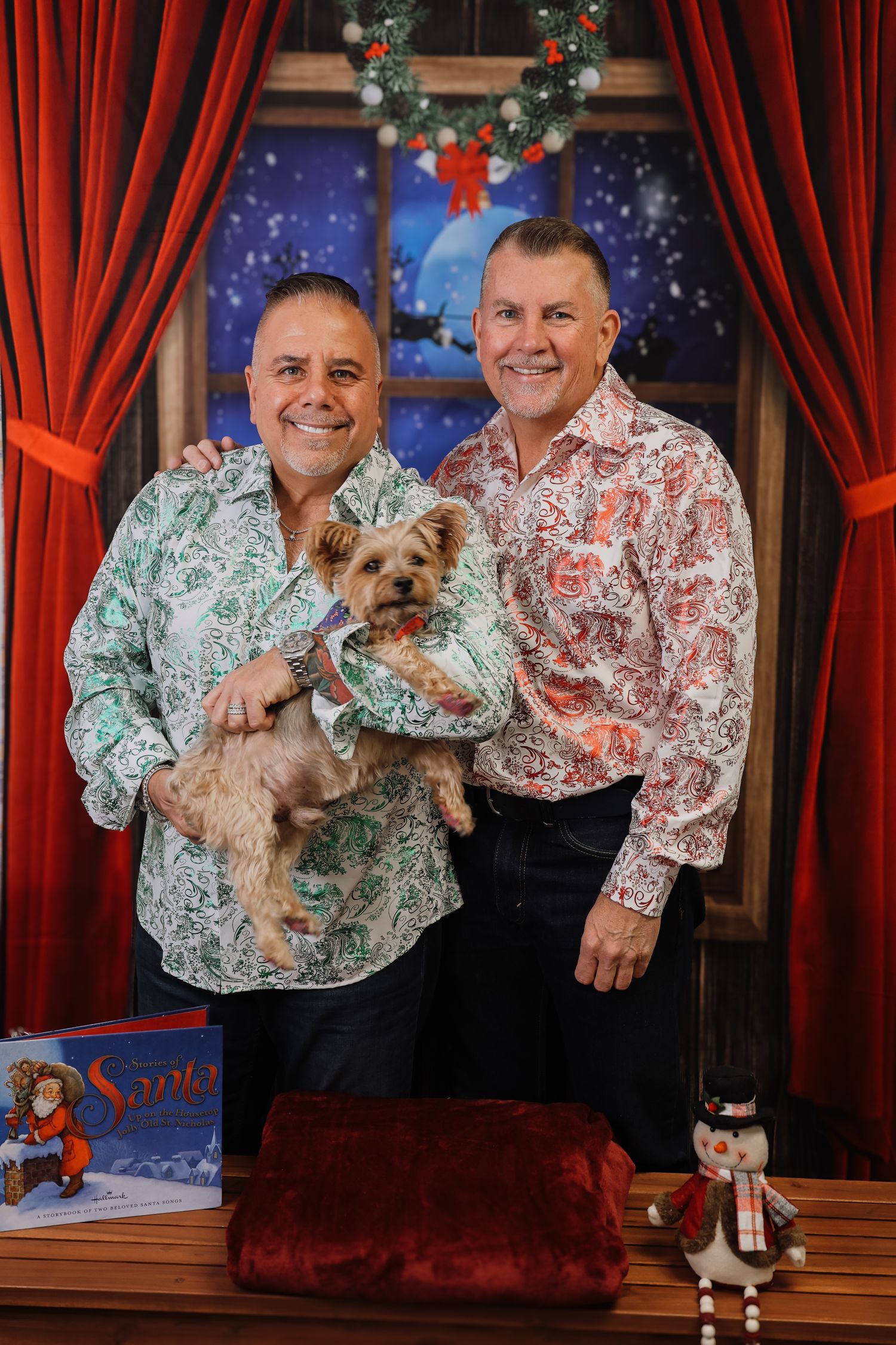 Two men in festive shirts pose with a Yorkshire Terrier in a Christmas-themed photo studio setting.