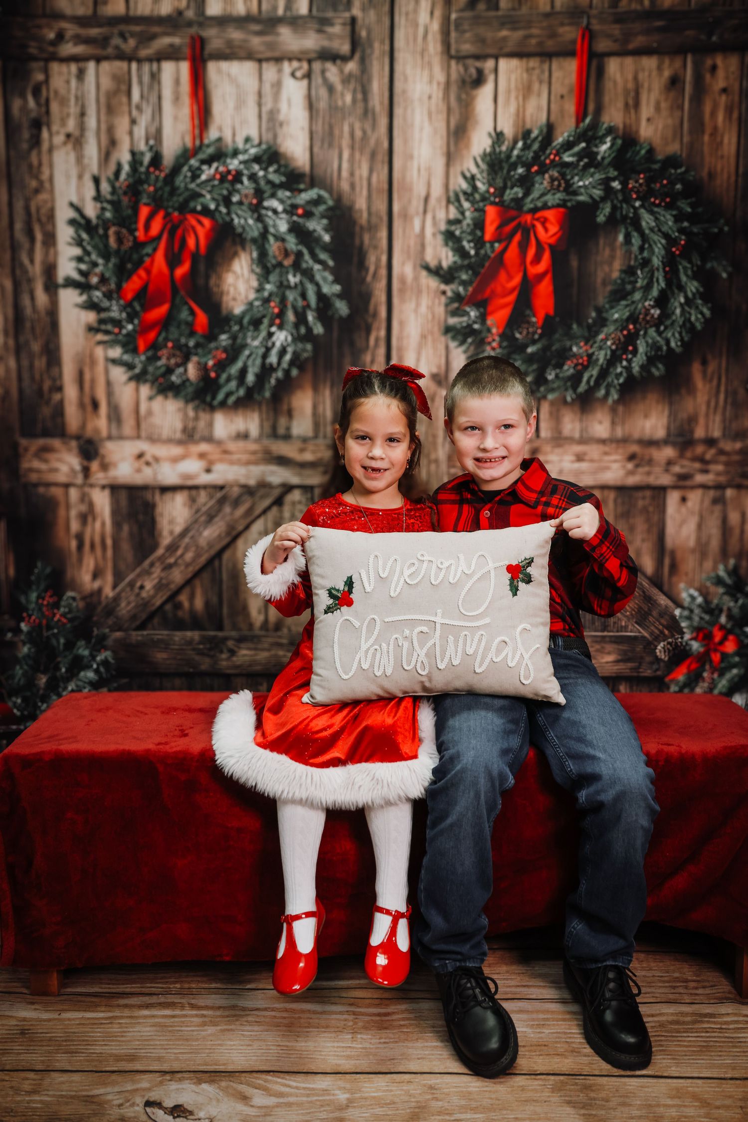 Two children in red Christmas outfits hold a Merry Christmas pillow in front of festive wreaths on a rustic wooden door.
