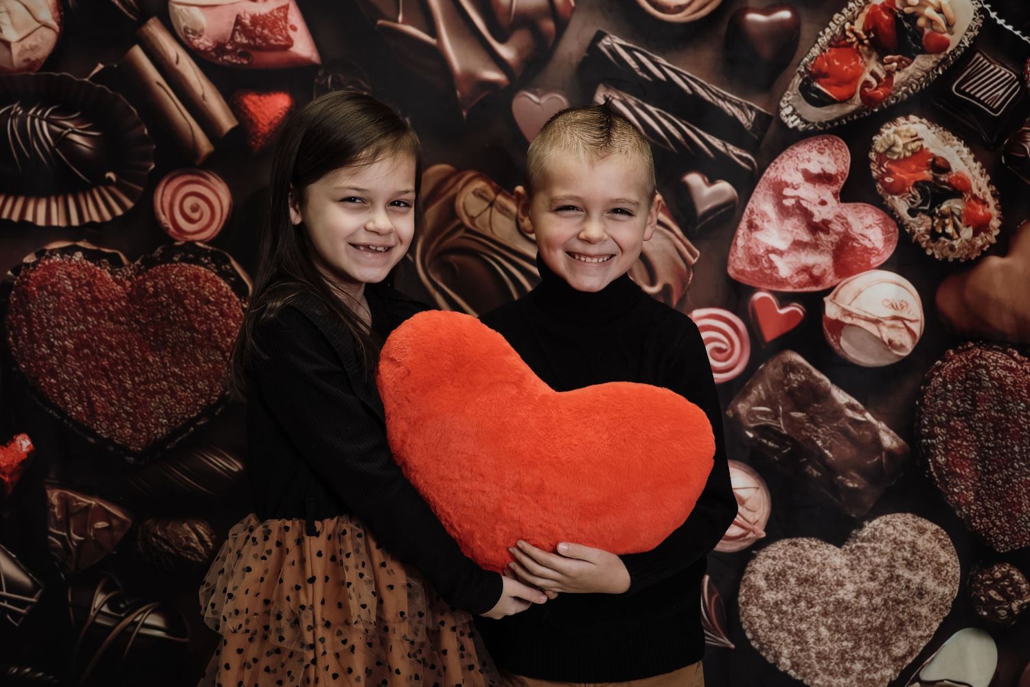 Two children smile holding a red heart pillow in front of a chocolate-themed Valentine's Day backdrop.