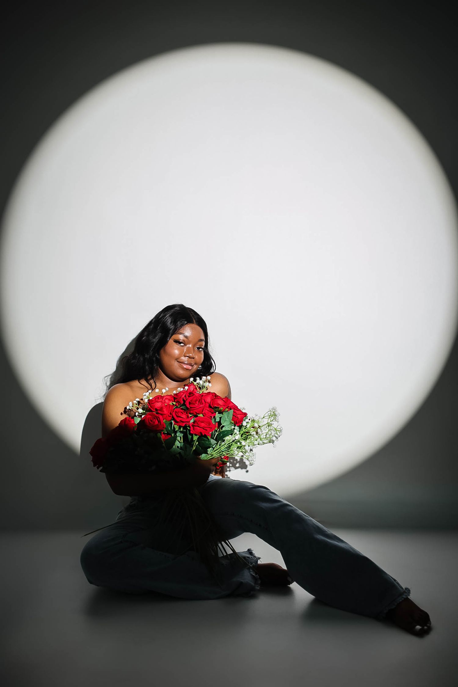 Woman sitting on floor holding large red rose bouquet in front of circular spotlight backdrop.