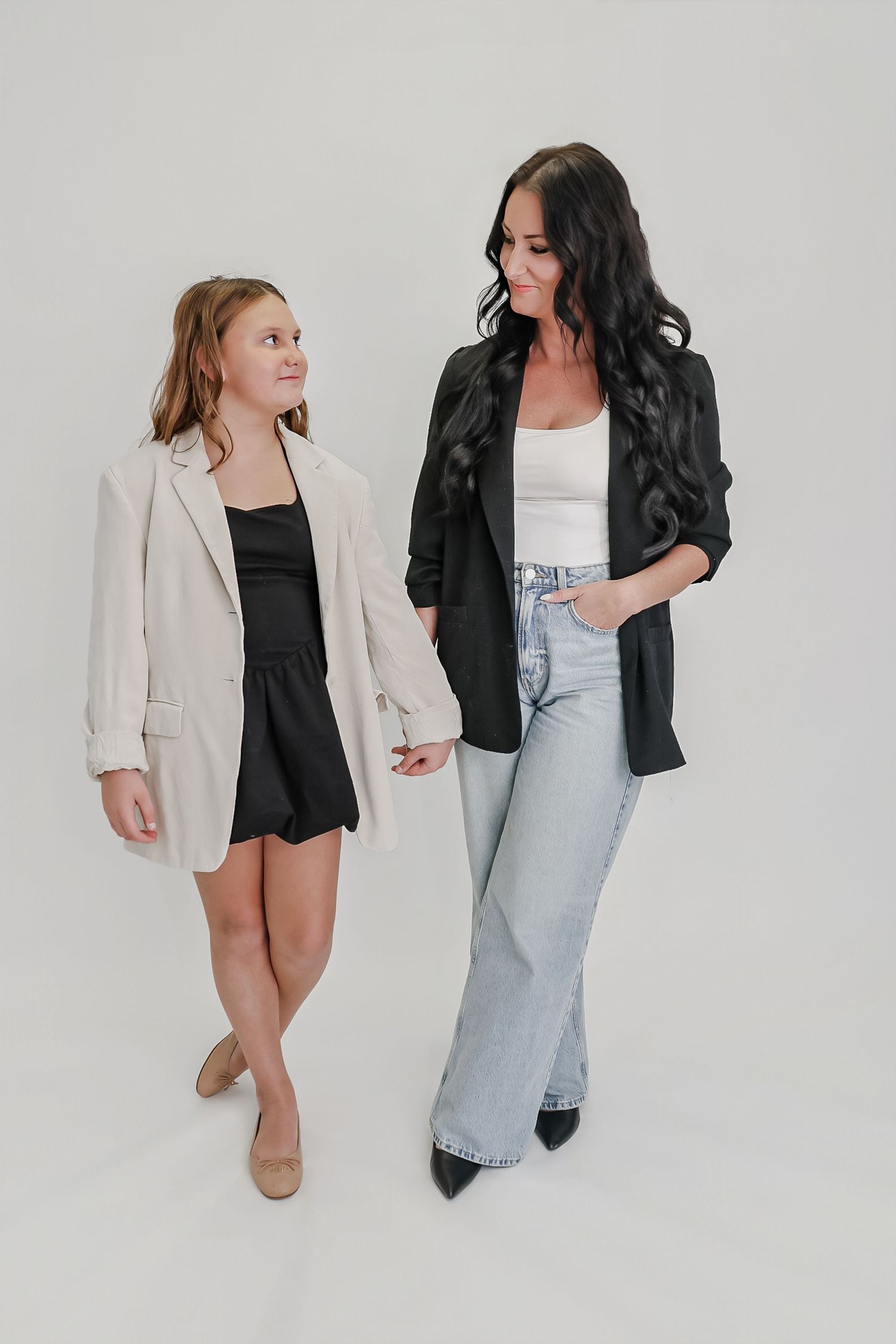 A woman and young girl in matching blazers and black outfits pose together against a white background.