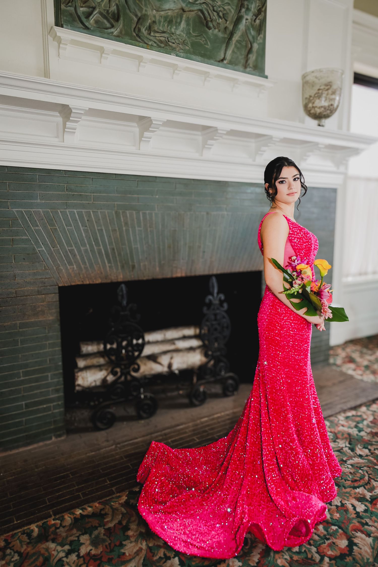 Woman in elegant red sequin gown holds colorful bouquet beside ornate fireplace in formal interior.