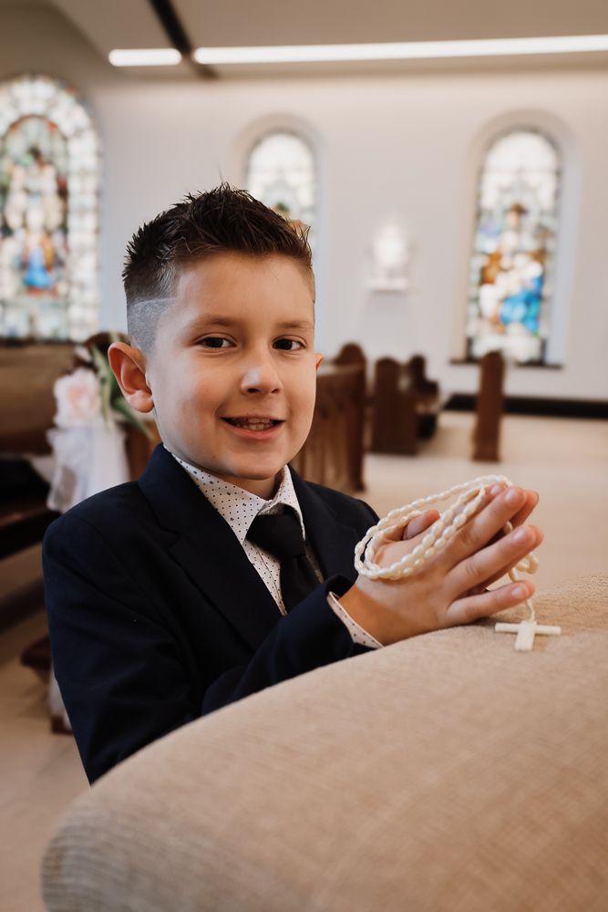Young boy in suit holding rosary beads in church pew during First Communion ceremony.