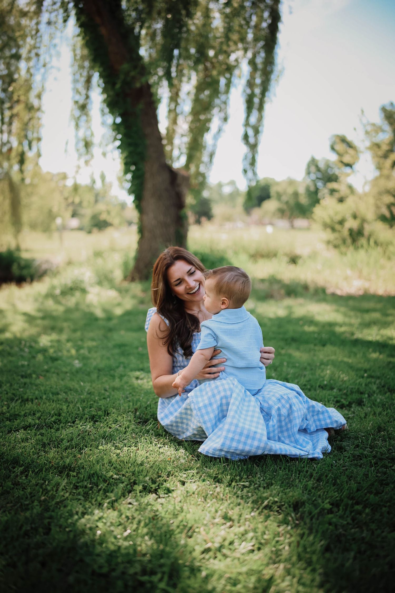 Smiling mother sitting on grass hugging adorable baby boy under willow tree in summer park.