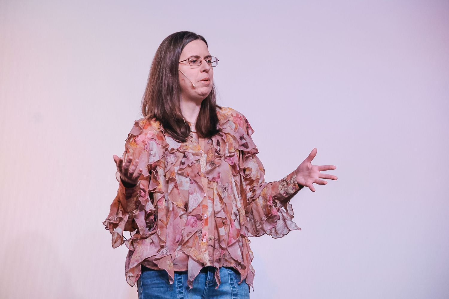 Woman in floral pink blouse and jeans gesturing with hand while speaking against light background.