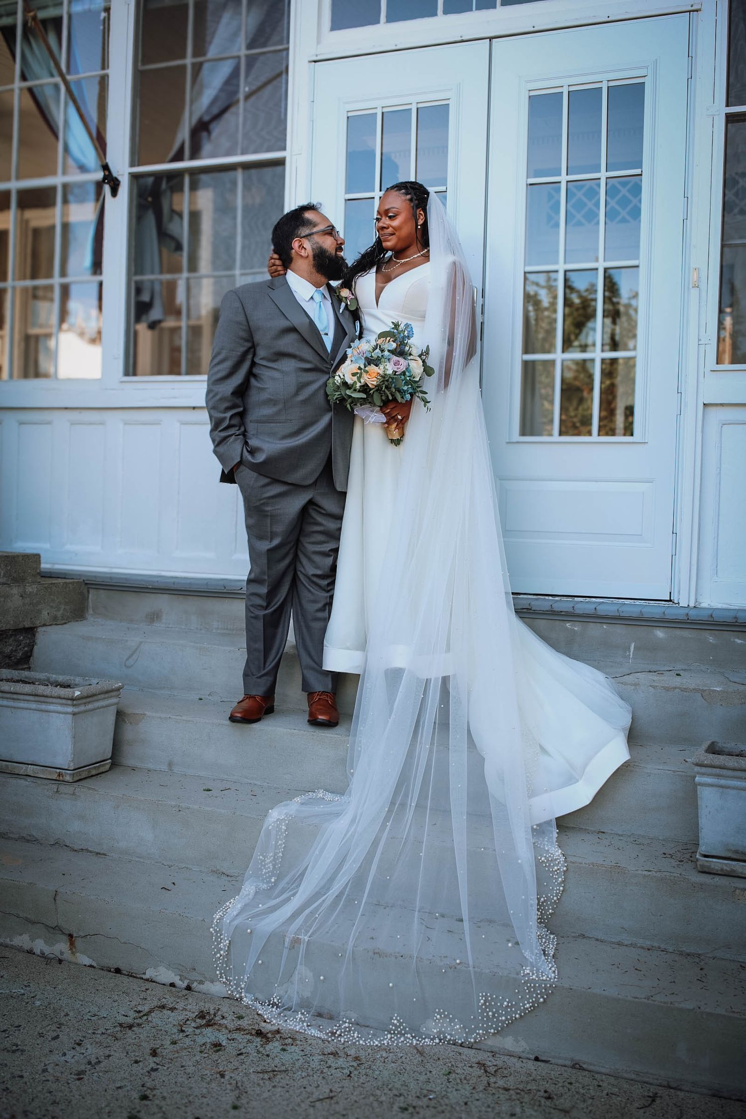 A bride in a flowing white gown with a long veil stands with her groom in a gray suit on elegant white steps.