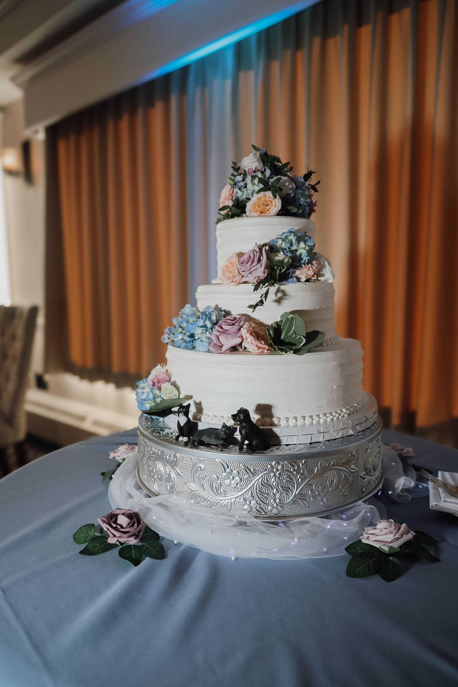 Elegant four-tier white wedding cake with pastel floral decorations on a silver stand, set against draped curtains.