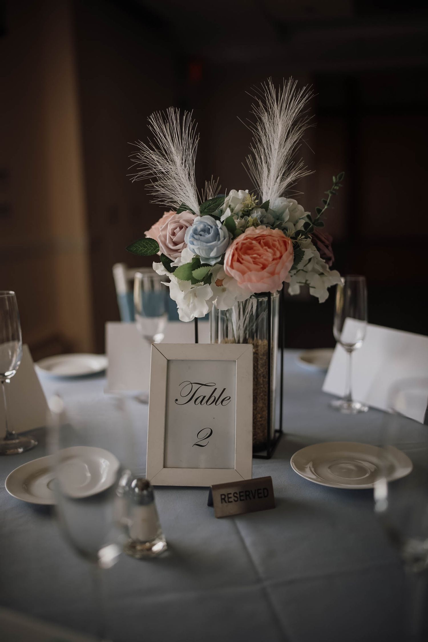 Elegant wedding table centerpiece with pink and blue roses, white feathers, and a framed Table 2 sign on a blue tablecloth.