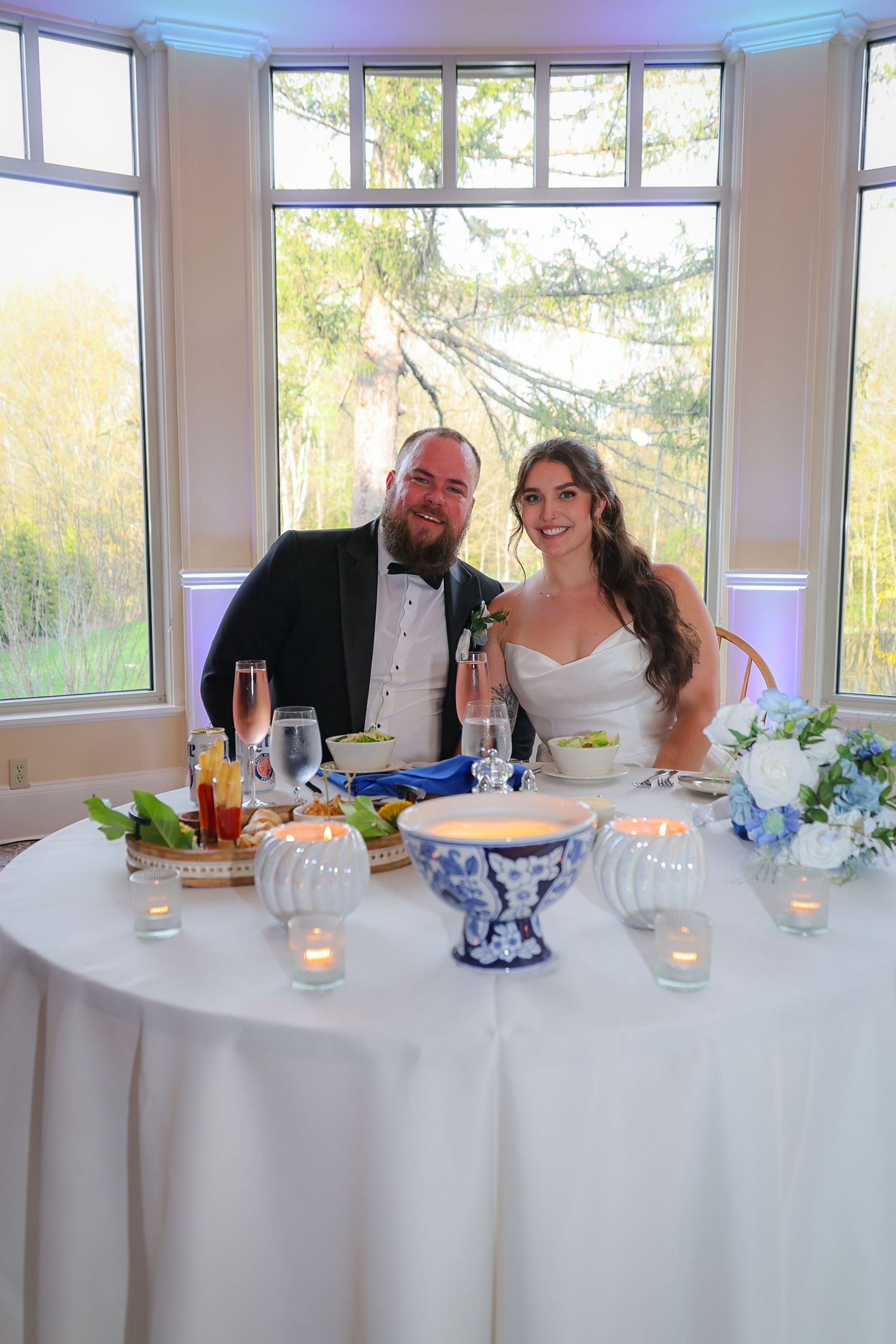 Bride and groom smile together at sweetheart table decorated with candles and blue floral arrangements at wedding reception.