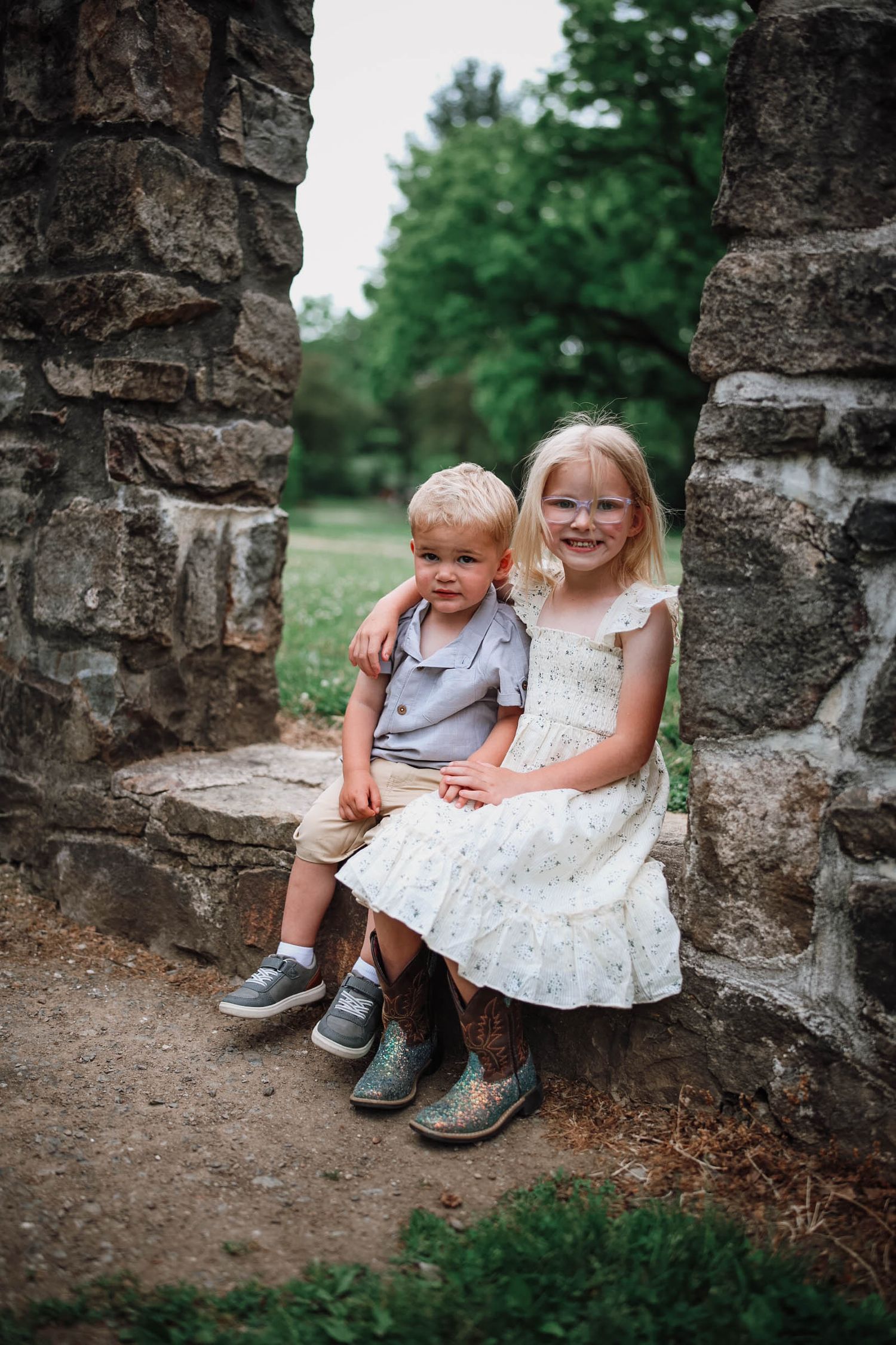 Two young siblings, a boy and girl, sit together on stone ruins in a park, girl wearing glasses and white dress.