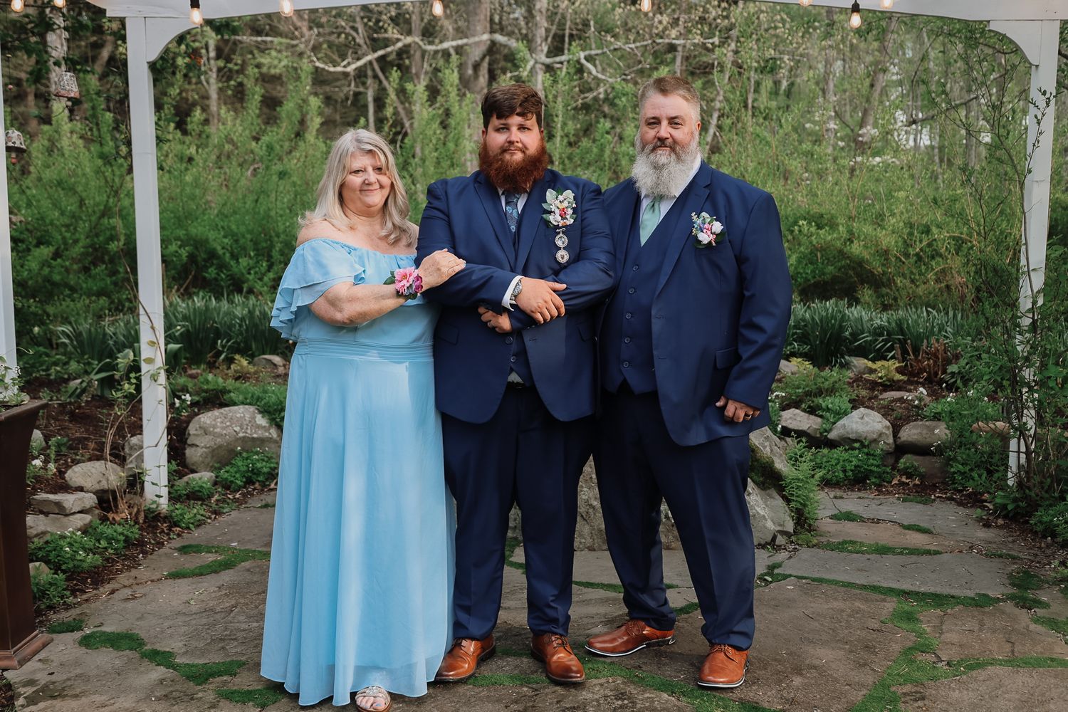 Three people in formal attire pose together in a garden wedding setting, two men in navy suits and a woman in a blue gown.
