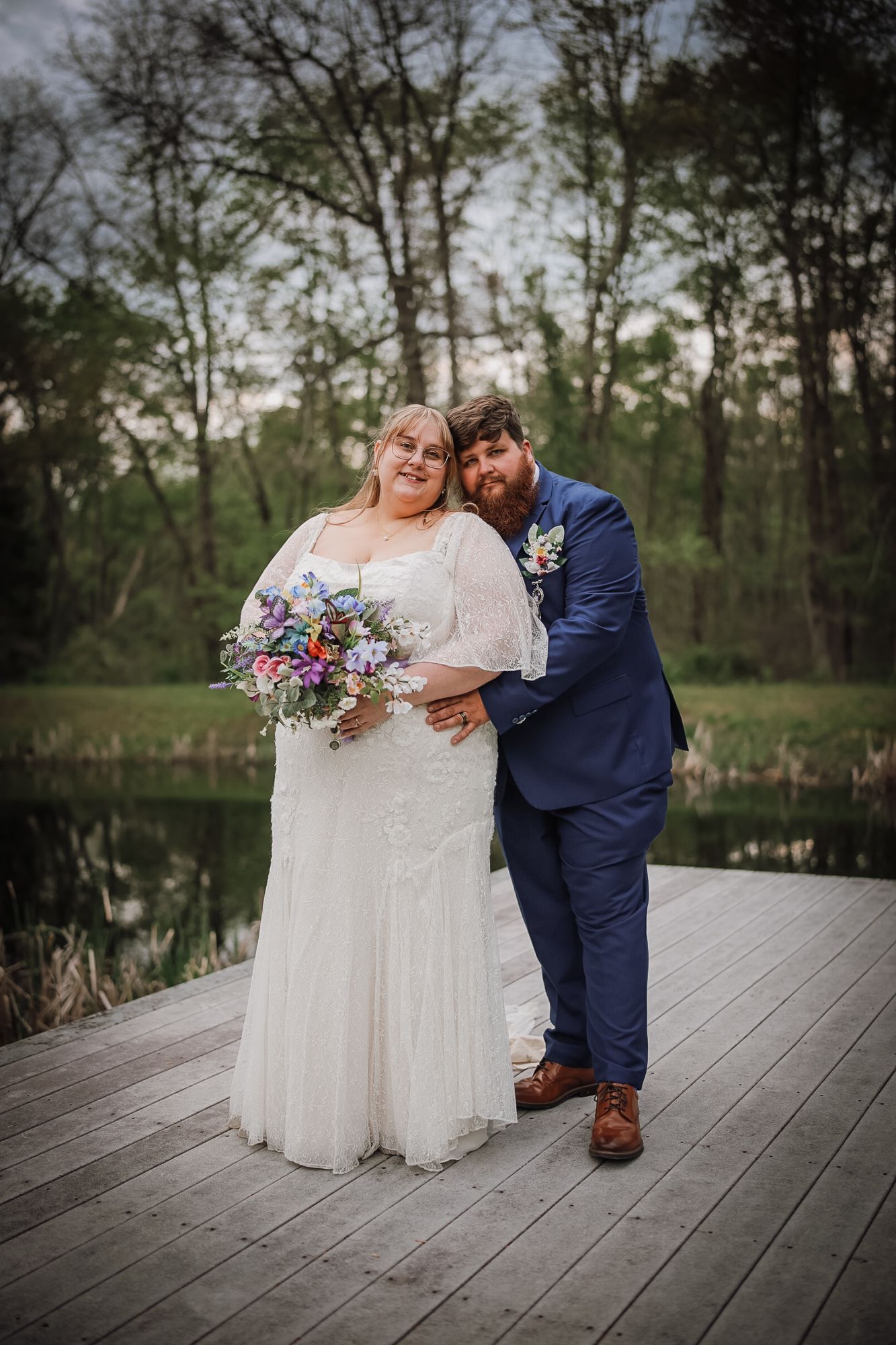 Bride and groom pose on wooden dock near pond, she holds colorful bouquet in white lace gown, he wears navy suit.