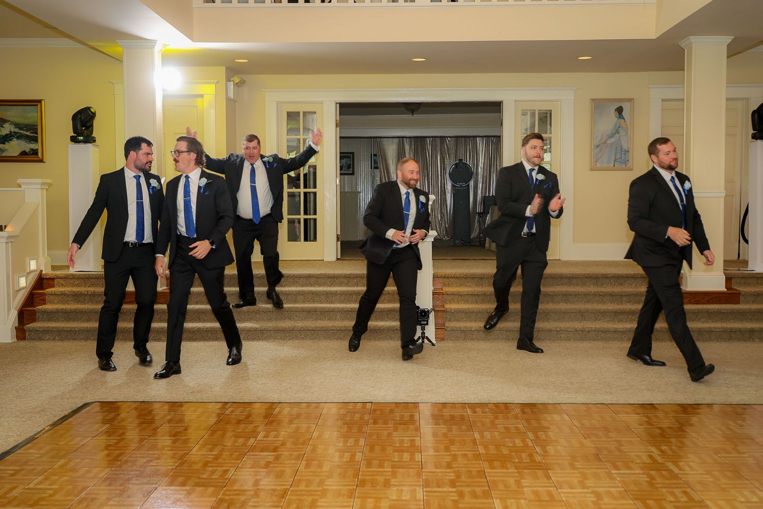 Groomsmen in black suits celebrate on a dance floor at an elegant wedding reception venue.