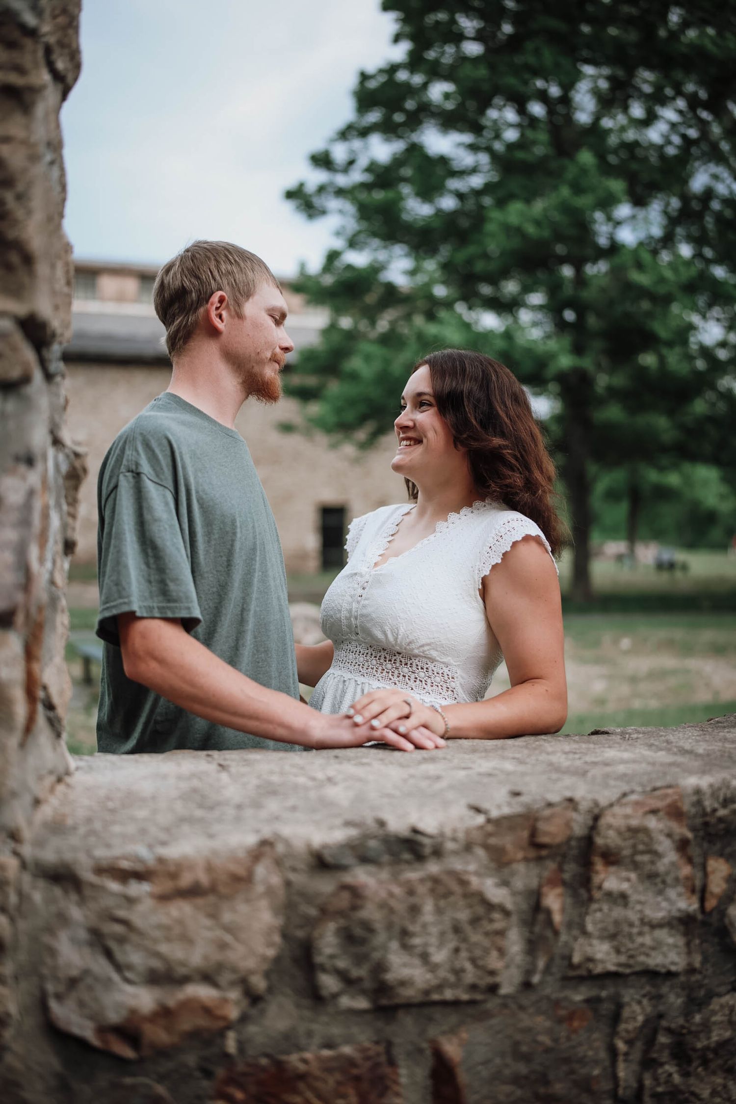 A young couple holds hands and gazes at each other near a stone wall outdoors, surrounded by lush green trees.