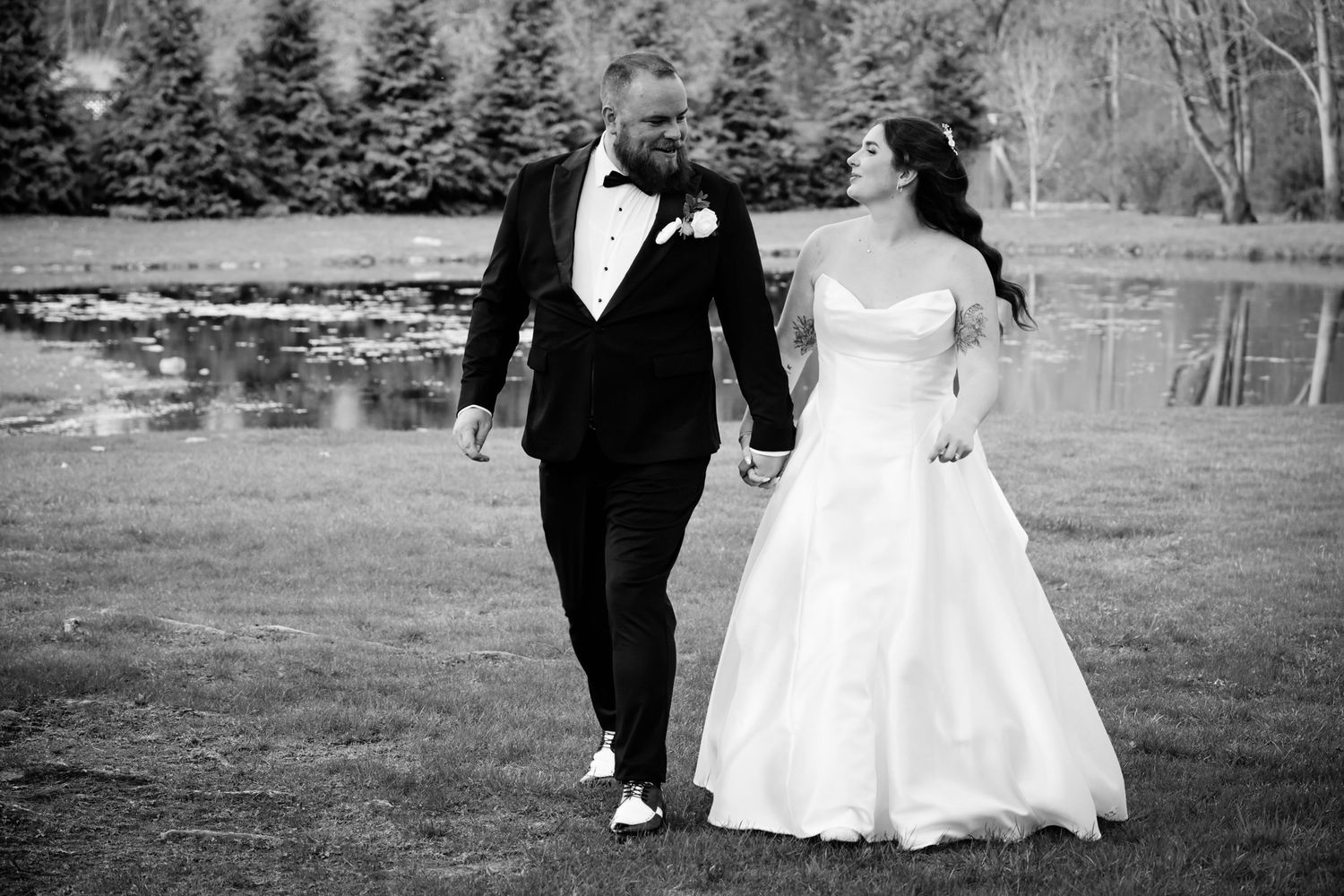 Black and white photo of a bride and groom holding hands walking outdoors near a pond on their wedding day.