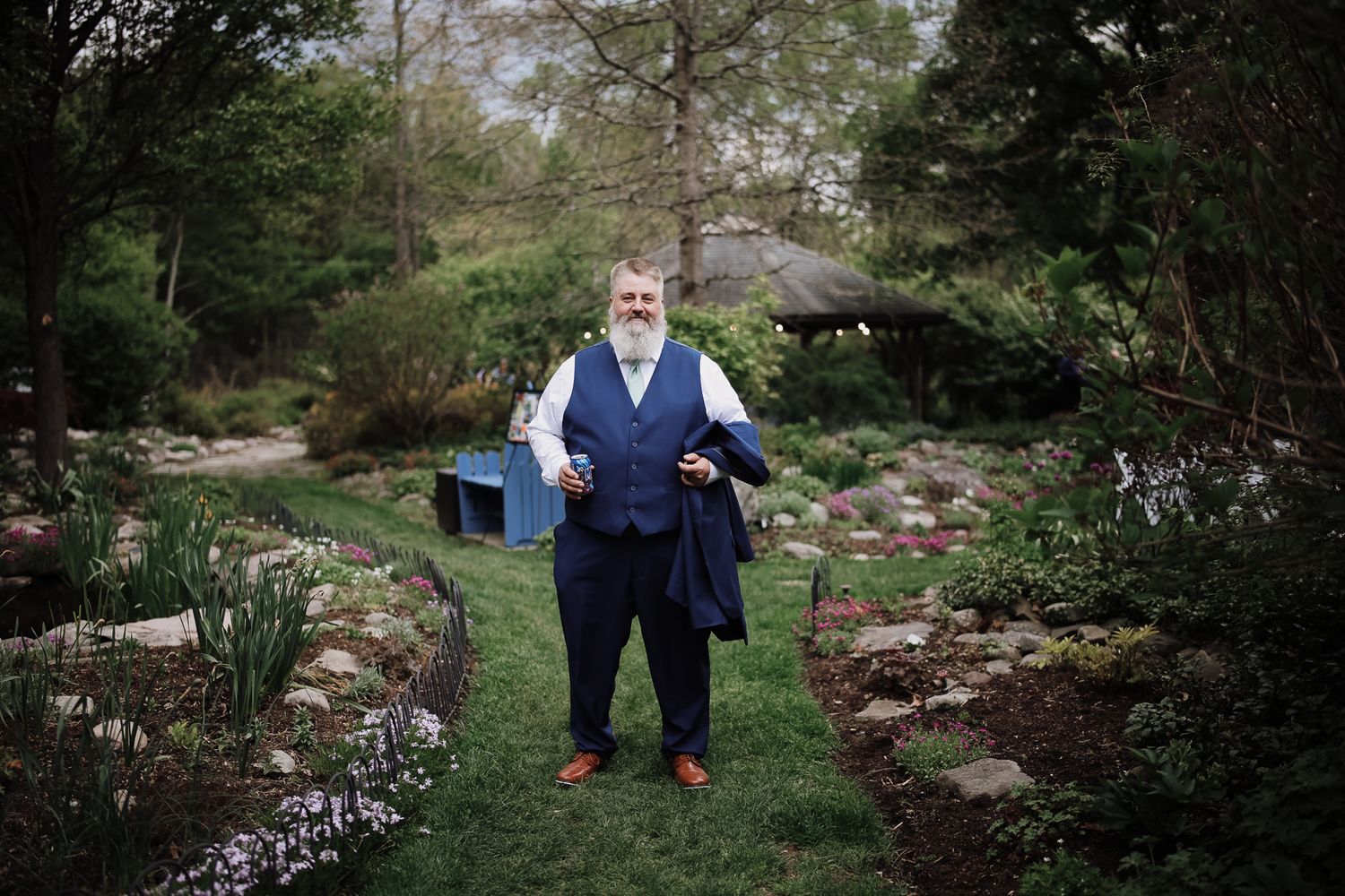 Bearded man in navy blue suit standing in a lush garden pathway, holding a jacket, surrounded by greenery.