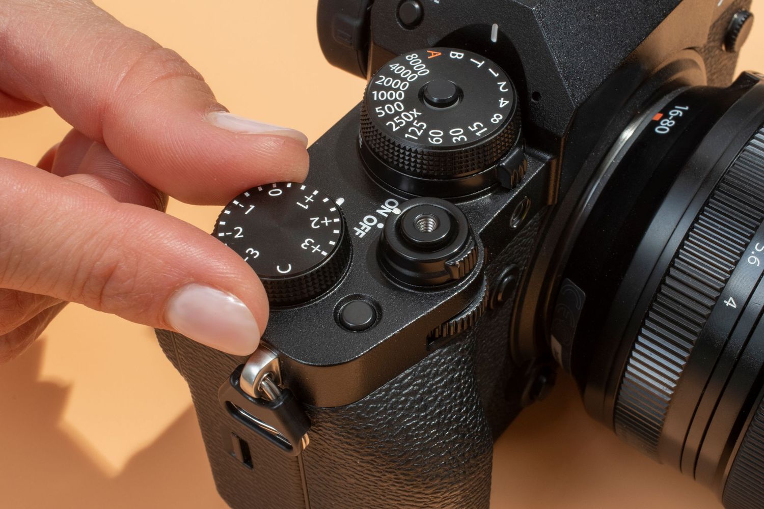 Fingers adjusting camera settings on a black digital camera with manual control dials against an orange background.