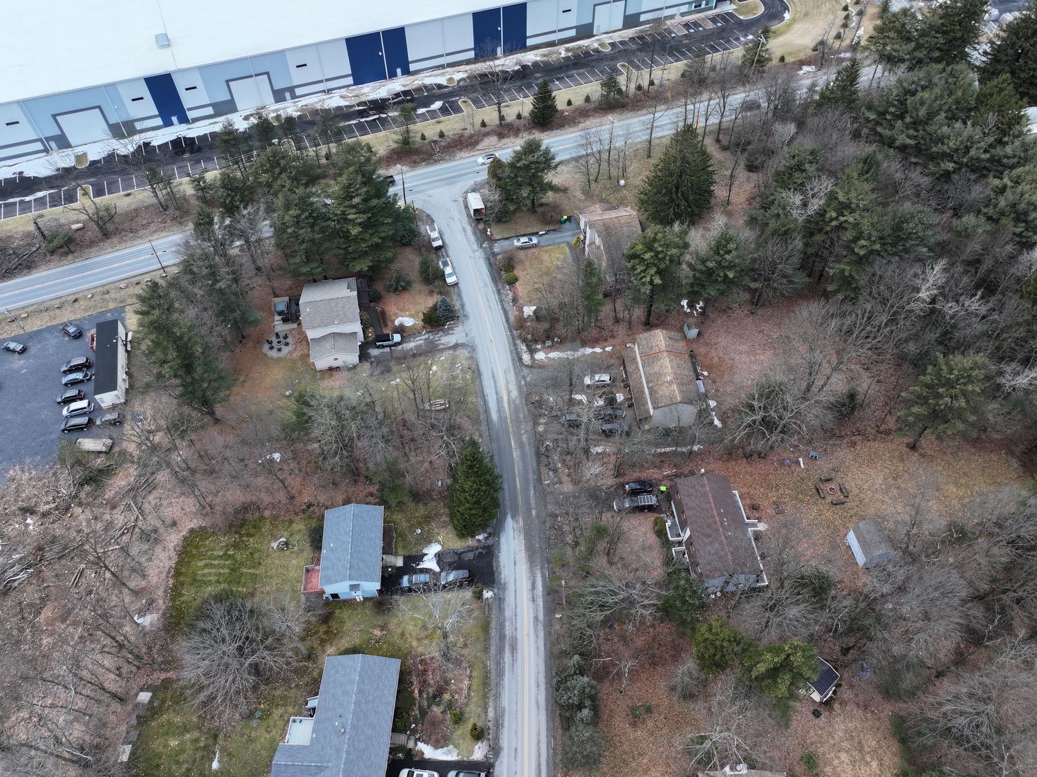 Aerial drone view of a rural road lined with bare trees and small properties near a large commercial warehouse building.