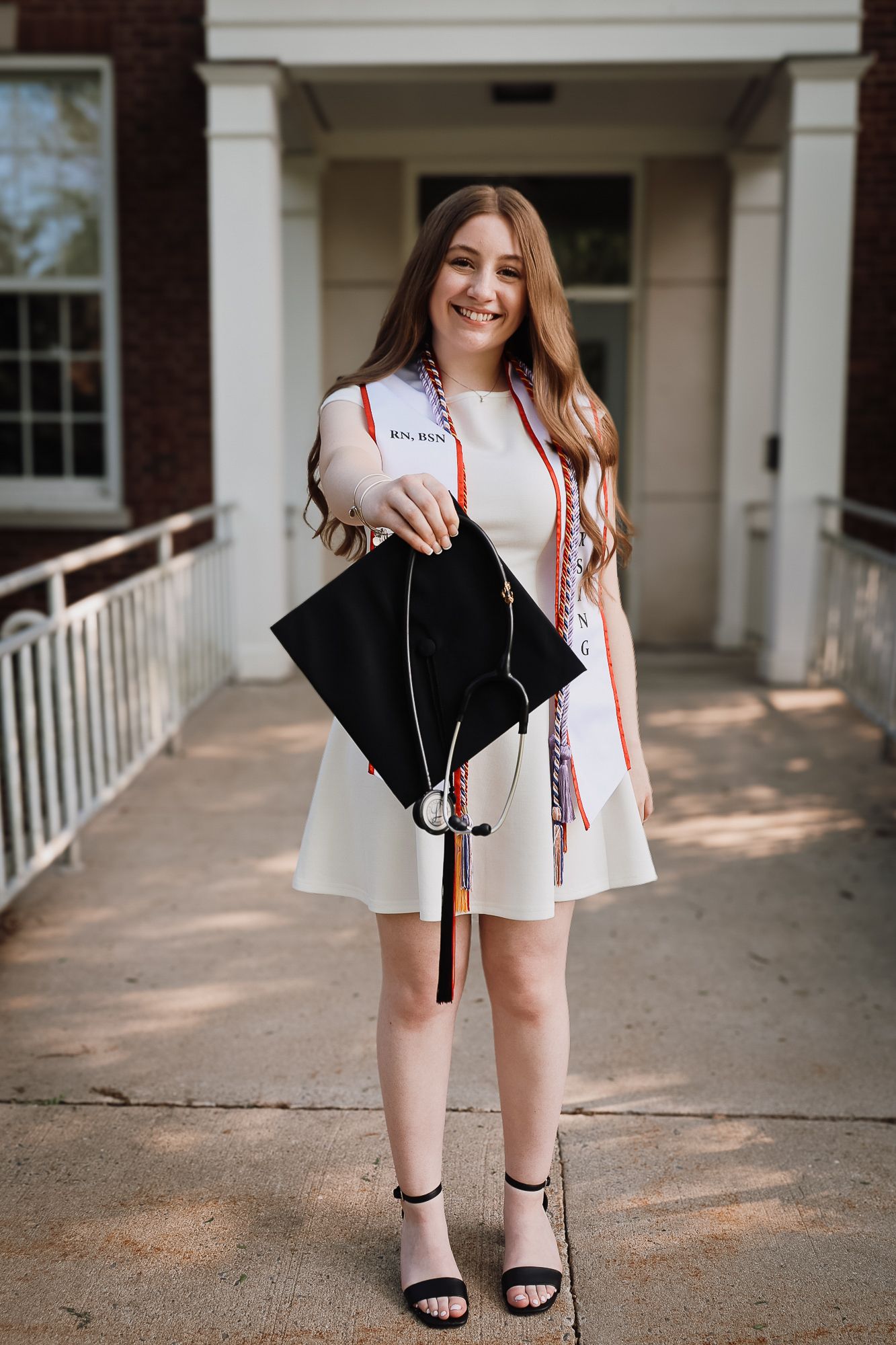Smiling graduate in white dress holding black cap with colorful cords and sash outside brick building.