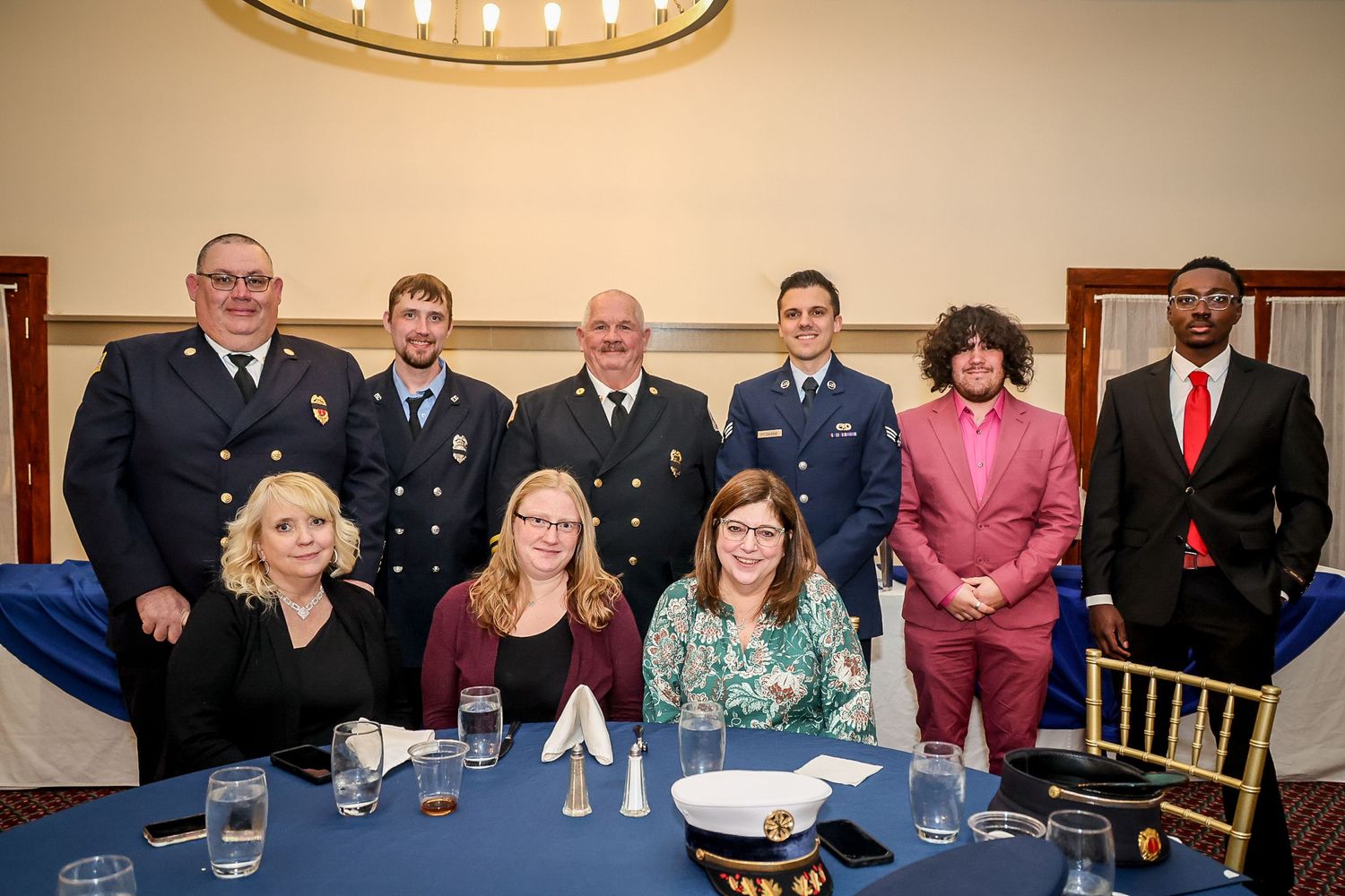 Group of nine professionals in formal attire and uniforms posing together at a banquet hall event.