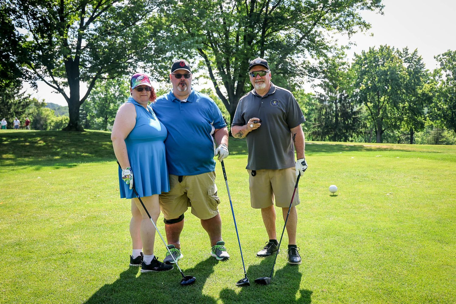 Three golfers posing on a sunny green golf course, holding clubs and smiling, surrounded by lush trees.