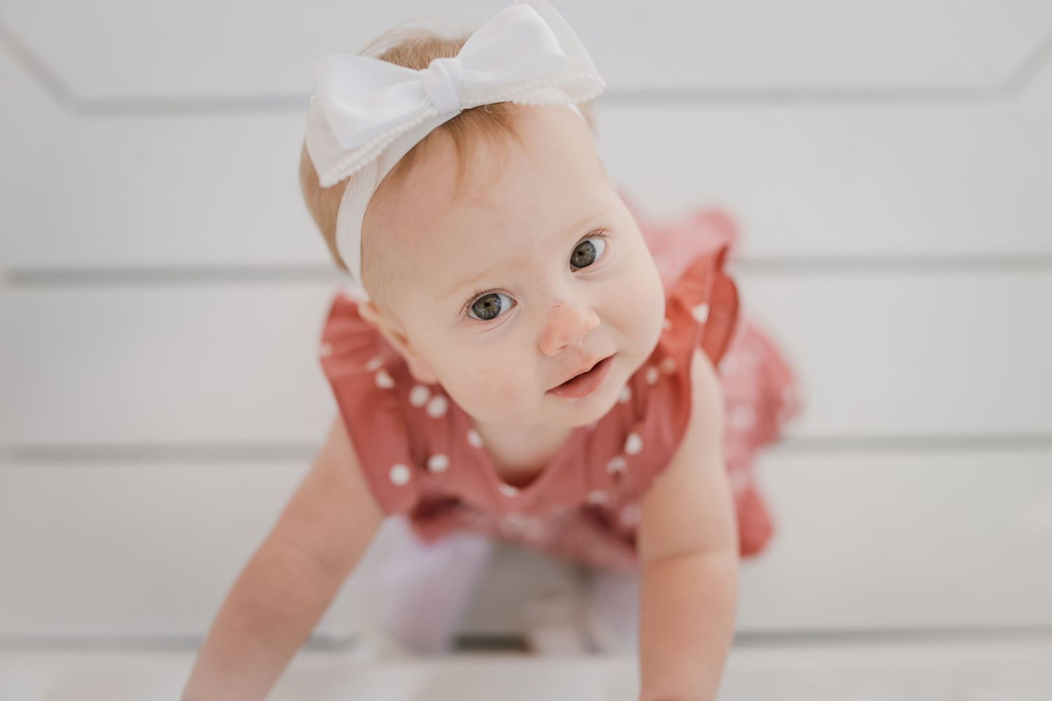 Adorable baby girl in pink ruffled outfit and white bow headband crawling on white floor.