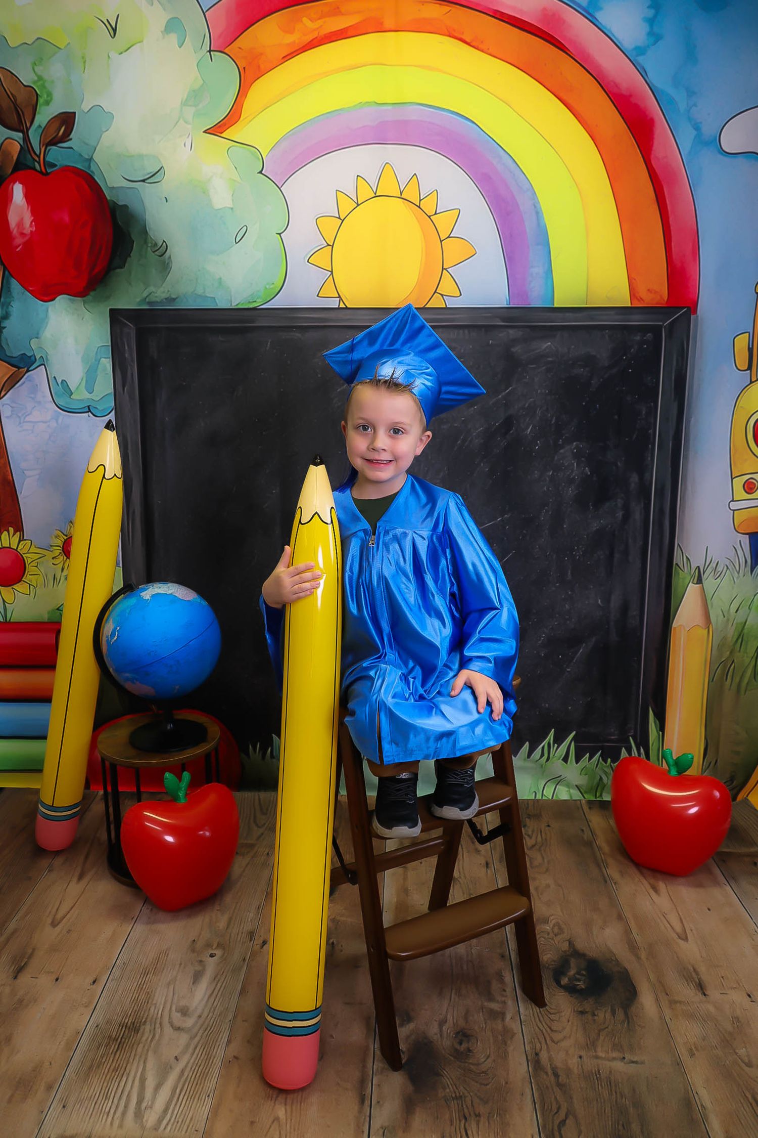 Young boy in blue graduation cap and gown poses with giant pencil in vibrant school-themed backdrop.
