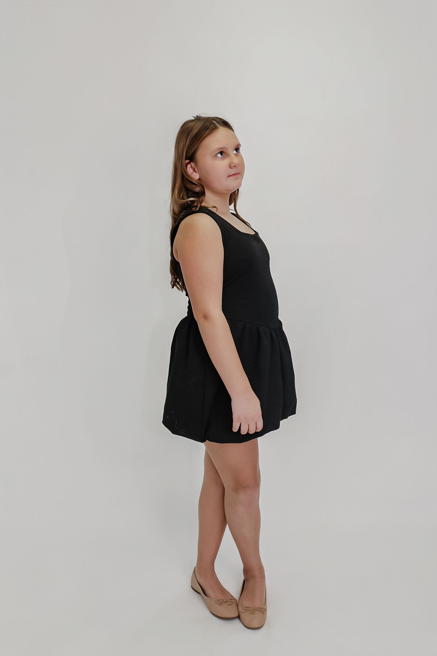A young girl poses in a black sleeveless mini dress with flared skirt and nude flats against a white background.
