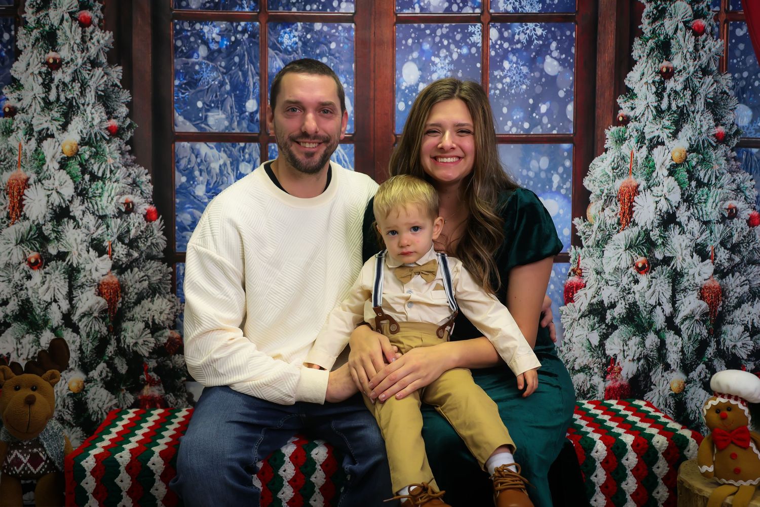 Family of three posing for a festive Christmas portrait with decorated trees and holiday backdrop.