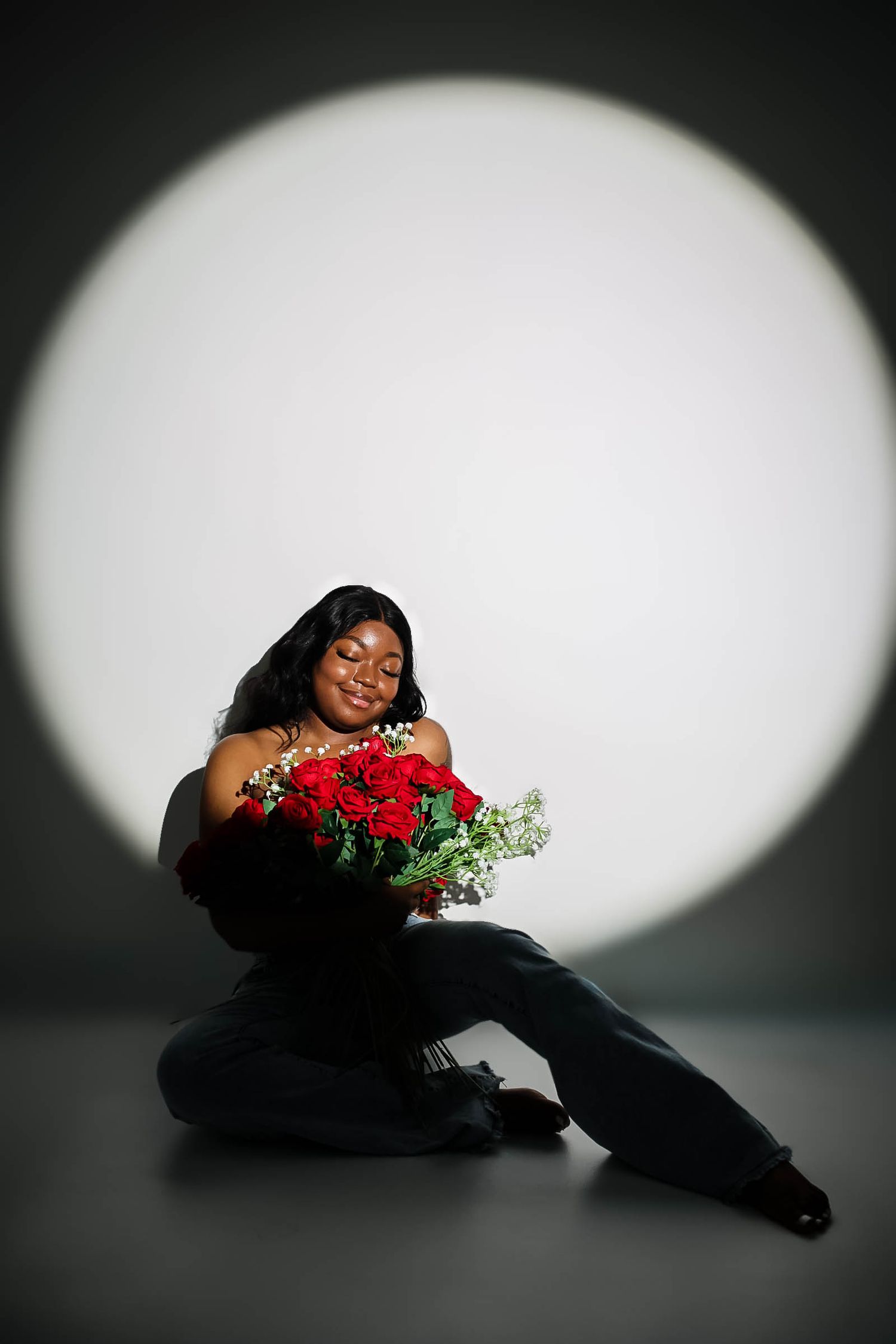 A woman sits on the floor holding a bouquet of red roses, illuminated by a circular spotlight against a dark background.
