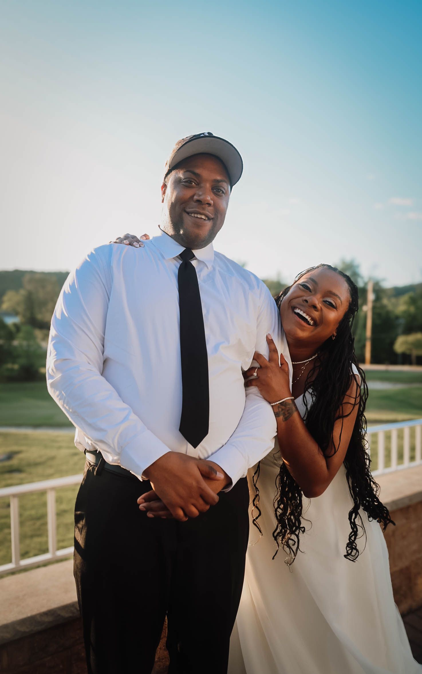 A smiling couple in formal attire posing outdoors at a sunny event venue.