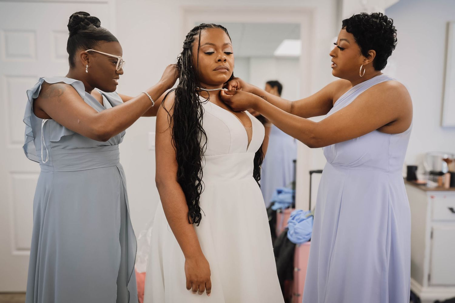 Two bridesmaids in light blue dresses help bride with her necklace during wedding day preparations indoors.