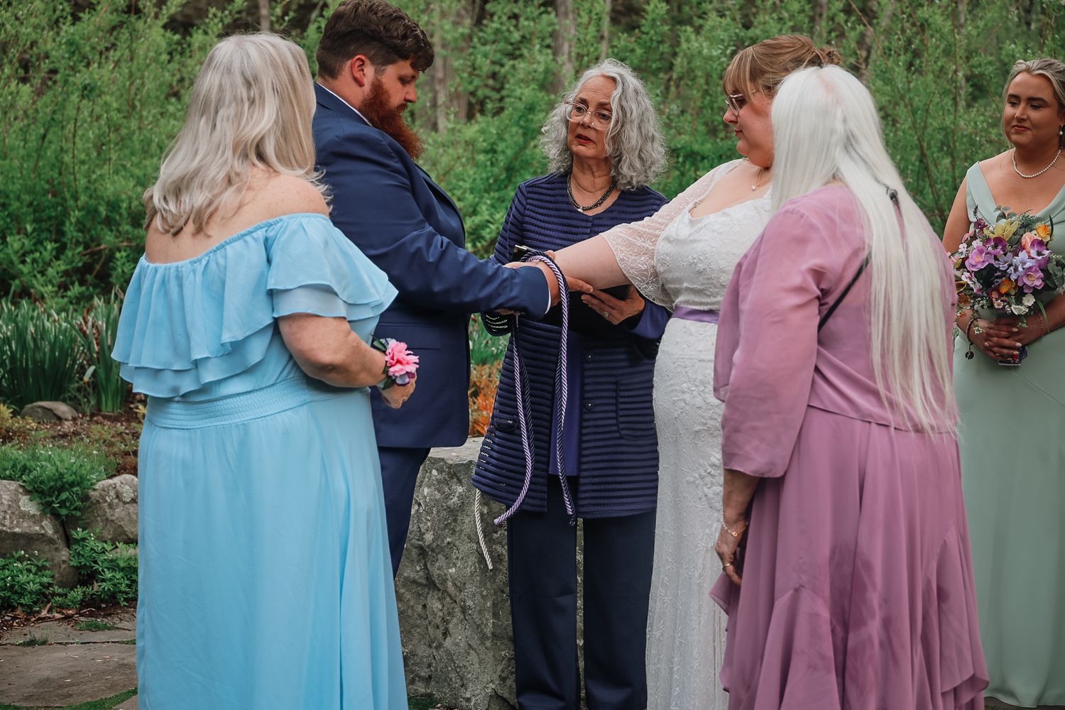 Outdoor wedding ceremony with couple exchanging vows, officiant, and bridesmaids in blue and purple dresses.
