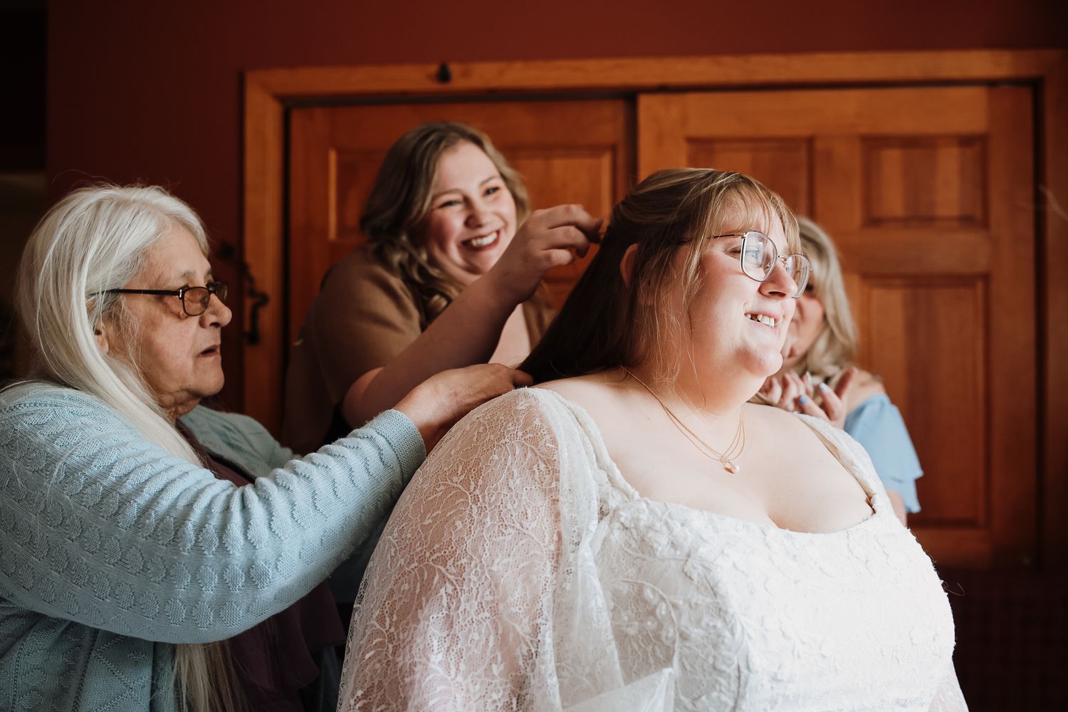 Bride in lace wedding dress getting ready as older woman and bridesmaid help with her hair, smiling warmly.