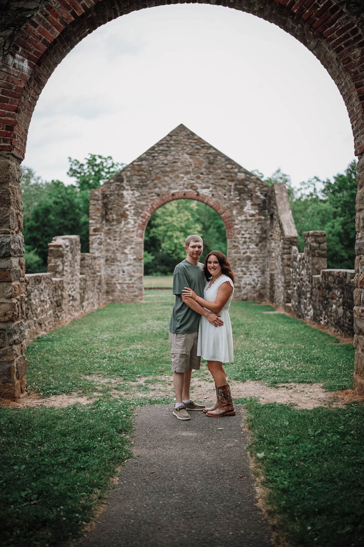 Couple embracing under brick archway at historic stone ruins, woman in white dress and boots, man in green shirt.