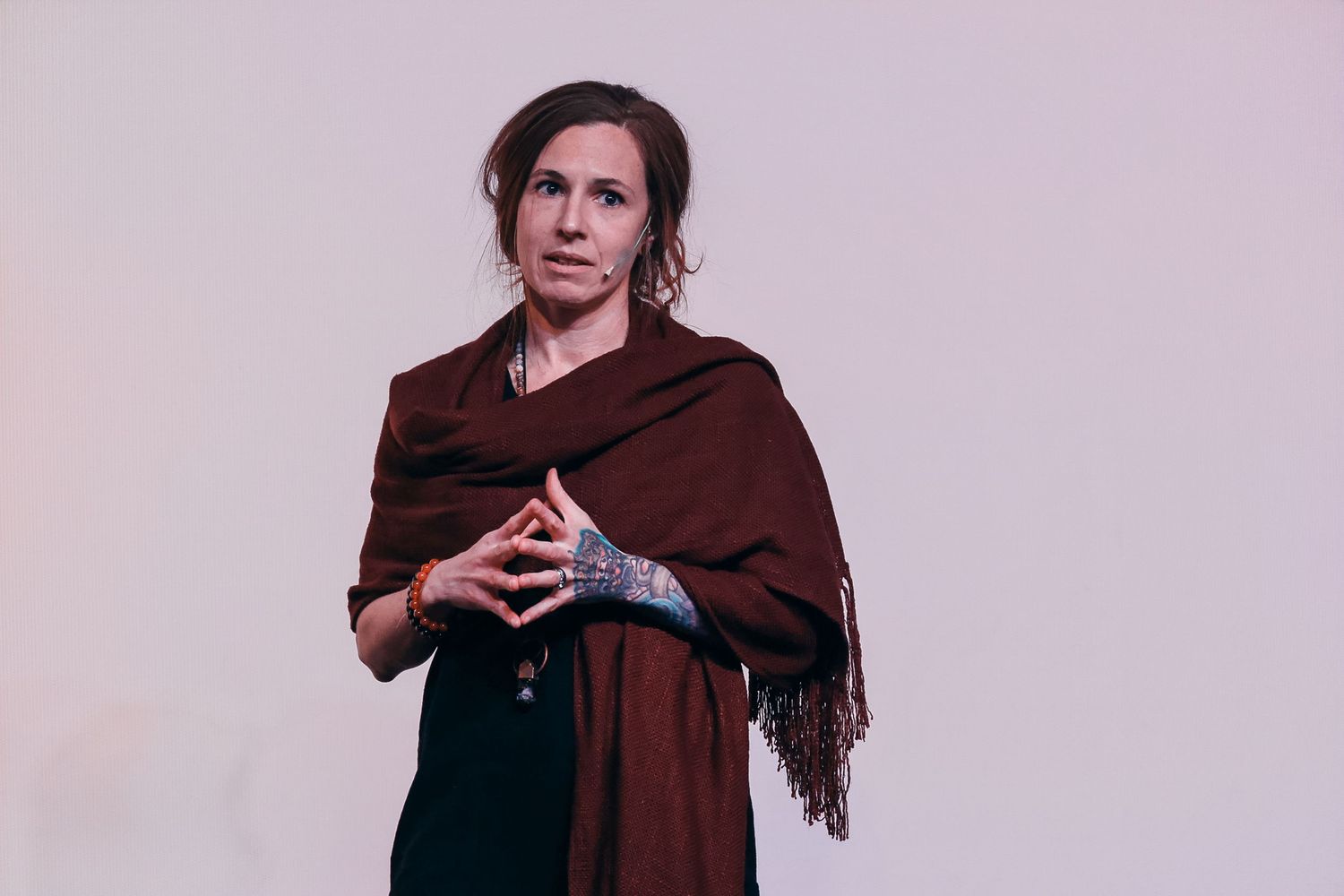 Woman in dark green dress and brown shawl with tattoed wrist stands against light background.