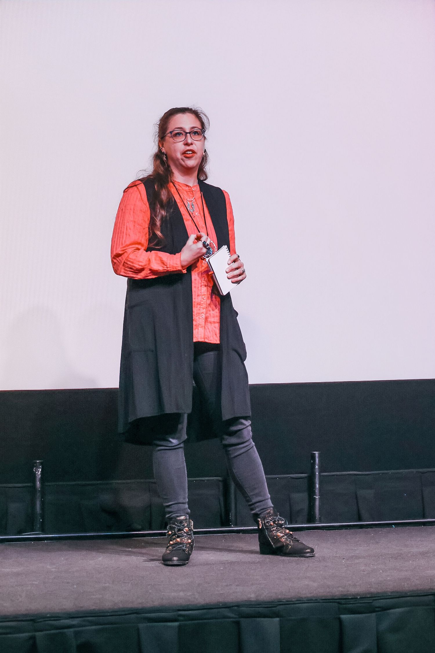 Woman in orange shirt and black vest speaking on stage at a conference event.