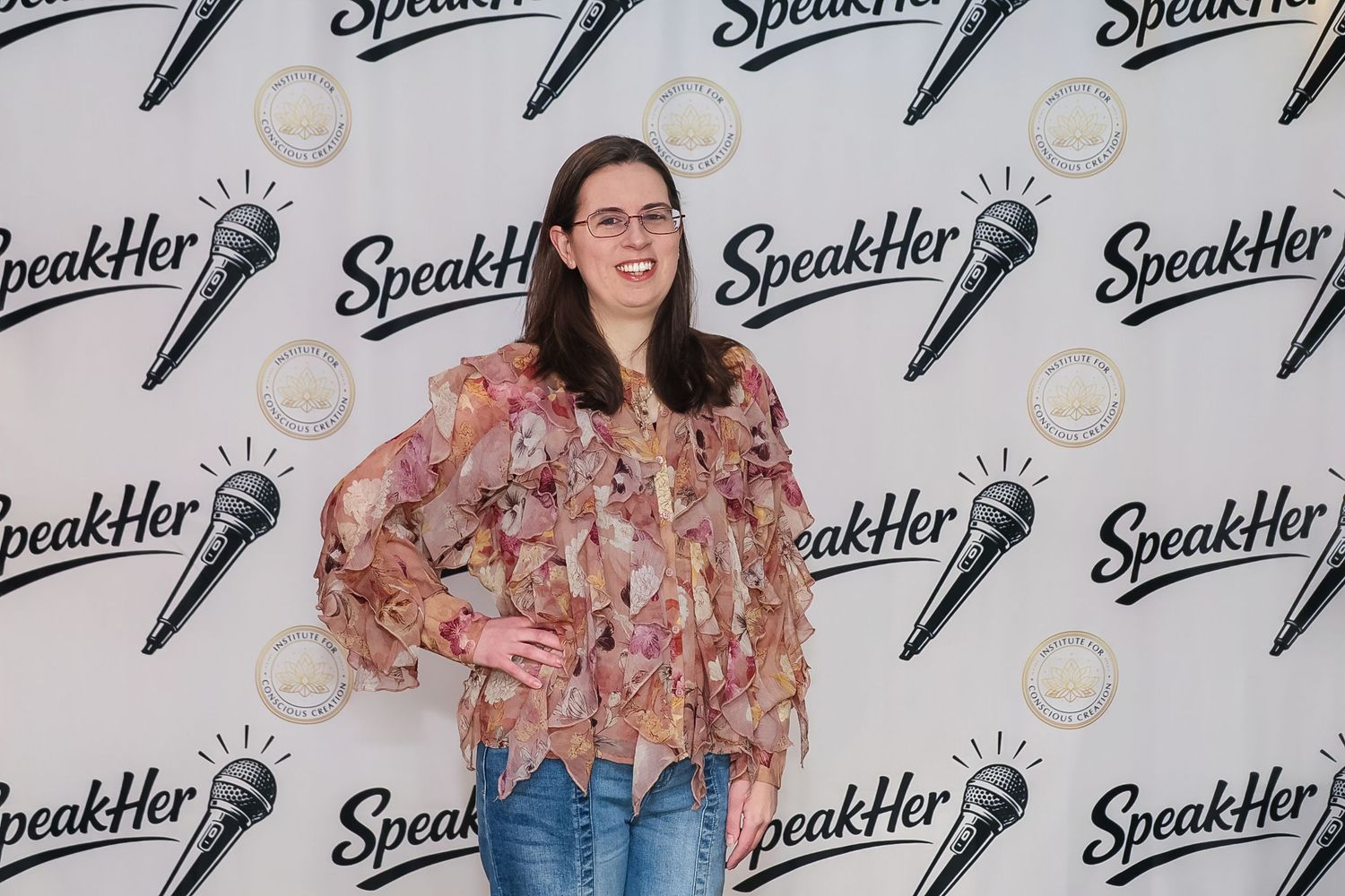 Woman in floral blouse smiling at SpeakHer event backdrop with microphone logos.