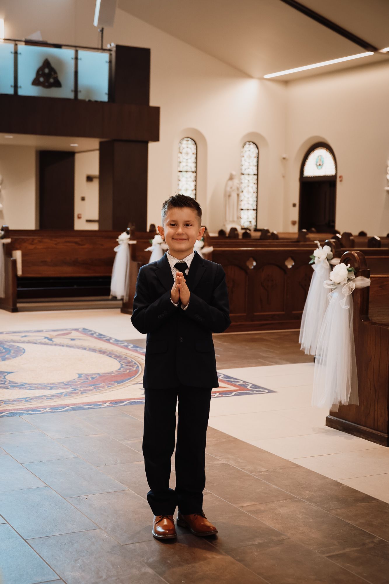 Young boy in black suit with hands clasped in prayer stance inside a decorated Catholic church.