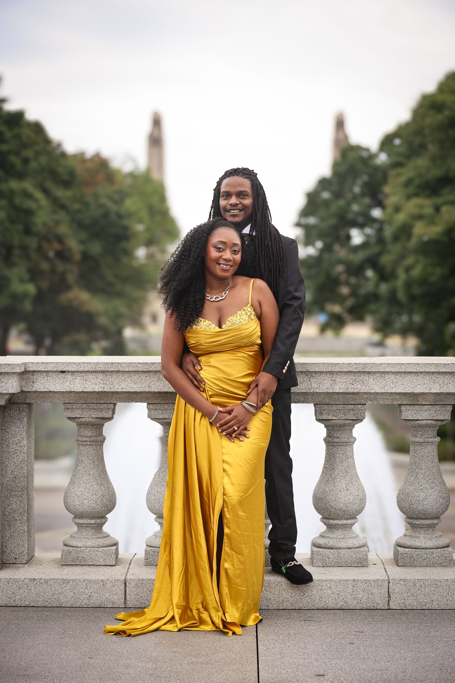 A couple in formal attire poses together on a stone balcony, with the person in yellow seated on the railing.