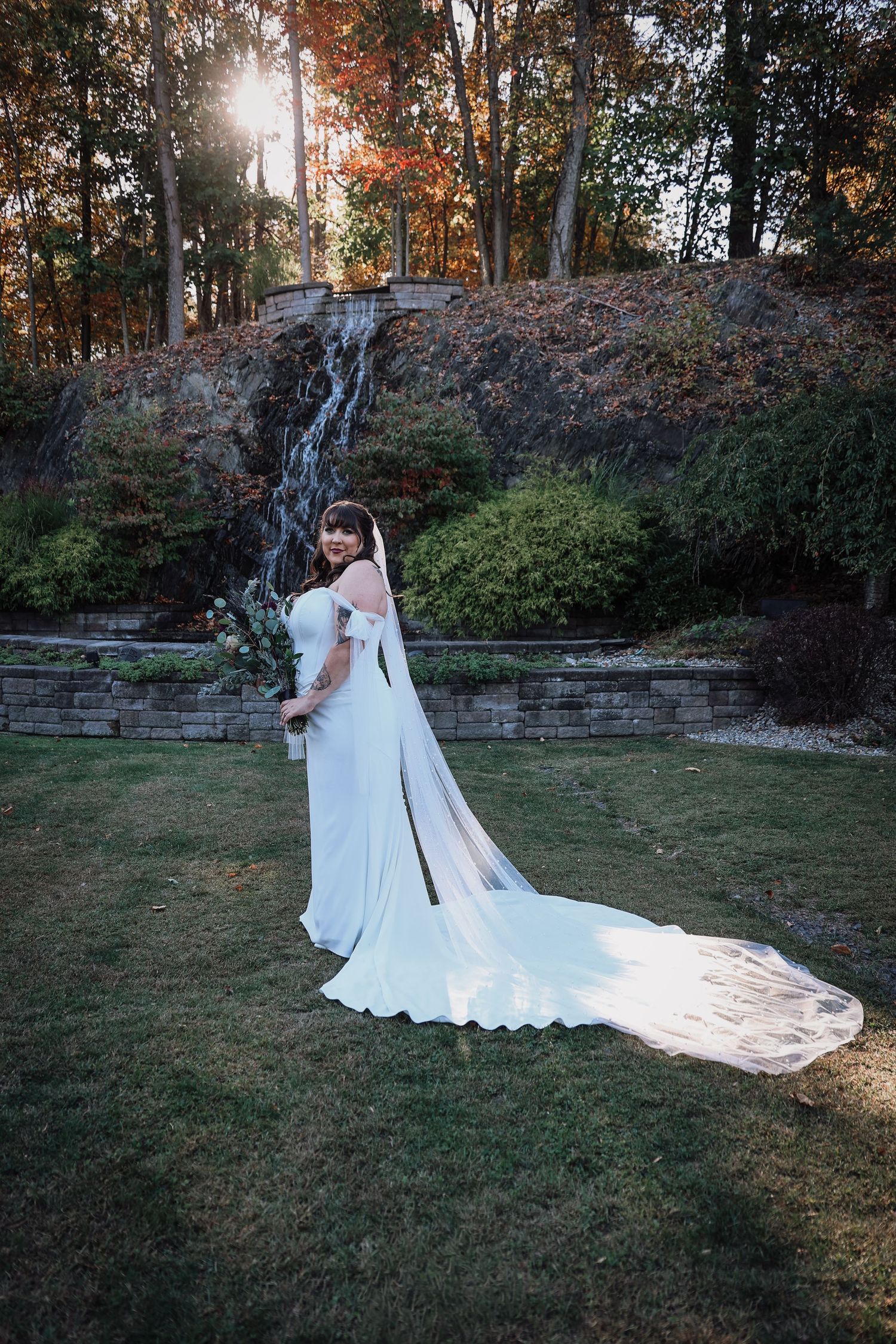 Romantic wedding couple embraces on a grassy lawn at sunset with stone wall and autumn trees in background.